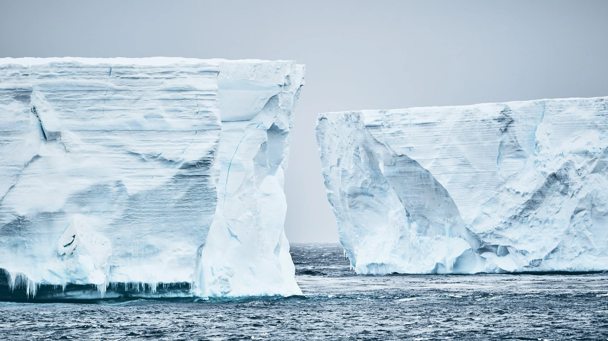 Two large icebergs floating in the ocean, with a gap between them revealing water underneath, under a gray sky.