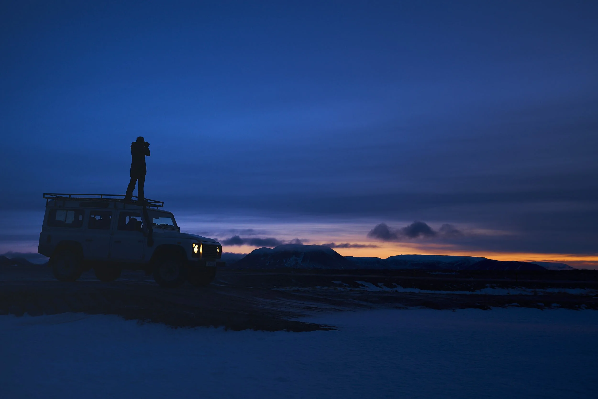 Silhouette of a person standing on top of a vehicle, looking through binoculars, against a sunset sky with mountains in the background.