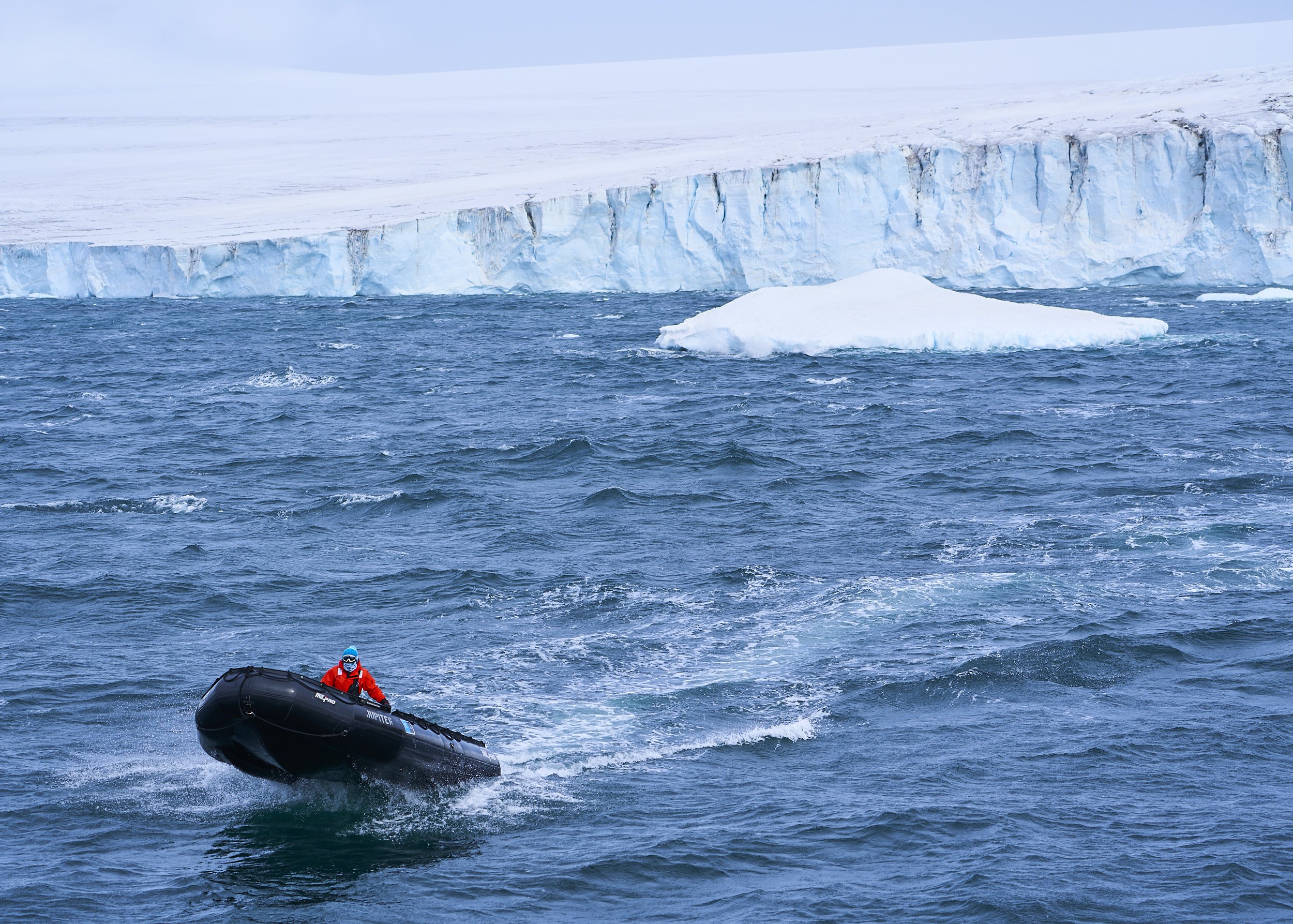 A person in a red jacket and blue helmet riding a black inflatable boat on icy blue ocean water, with a large ice cliff and floating ice in the background.