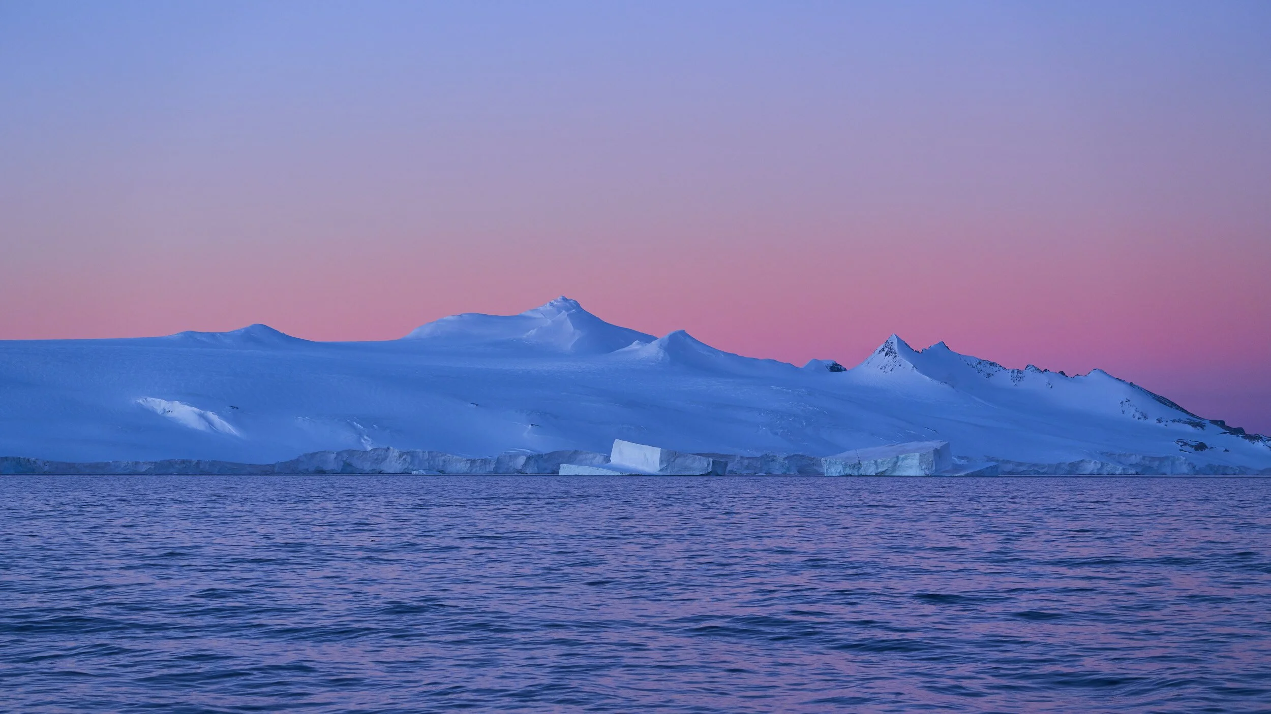 Iceberg floating in calm ocean with snow-covered mountain in the background under a clear sky at sunset or sunrise.