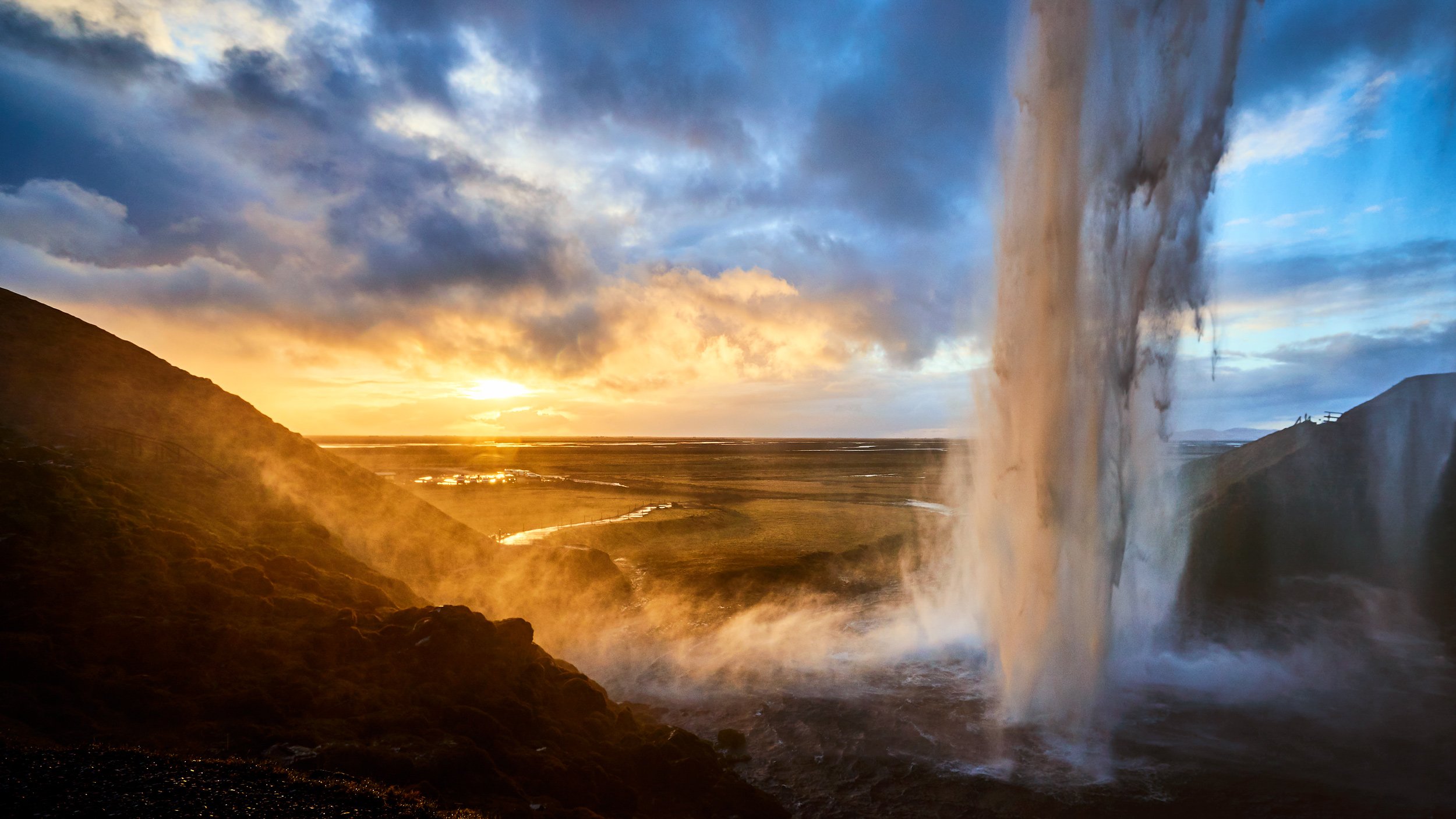 A scenic view of a waterfall at sunset, with clouds in the sky and a landscape of fields and hills in the background.