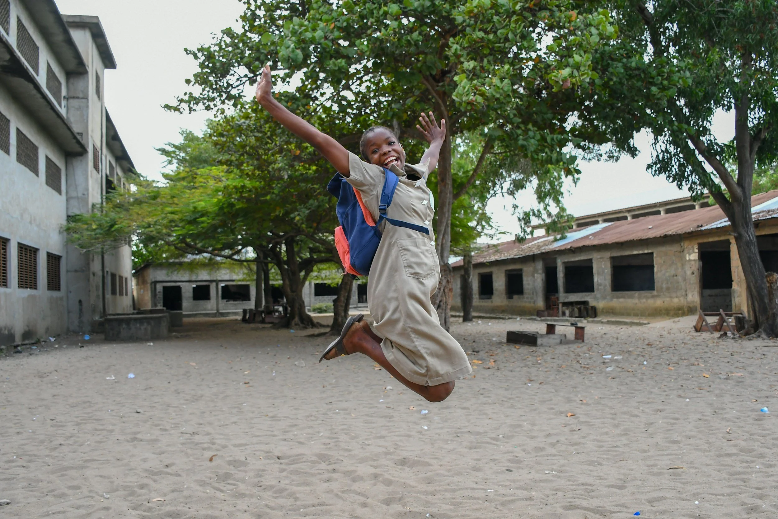 A young boy in a school uniform jumps joyfully on a dirt schoolyard with trees and buildings in the background.