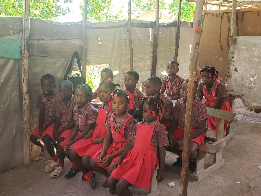 A group of young school children wearing school uniforms of red dresses and plaid shirts, seated on benches inside a classroom with makeshift wooden and fabric walls.