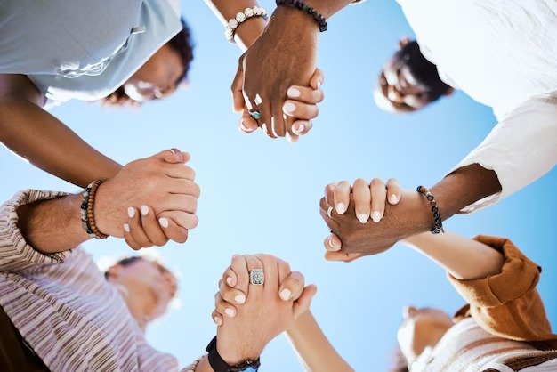 Group of diverse people holding hands in a circle, viewed from below with a blue sky background.