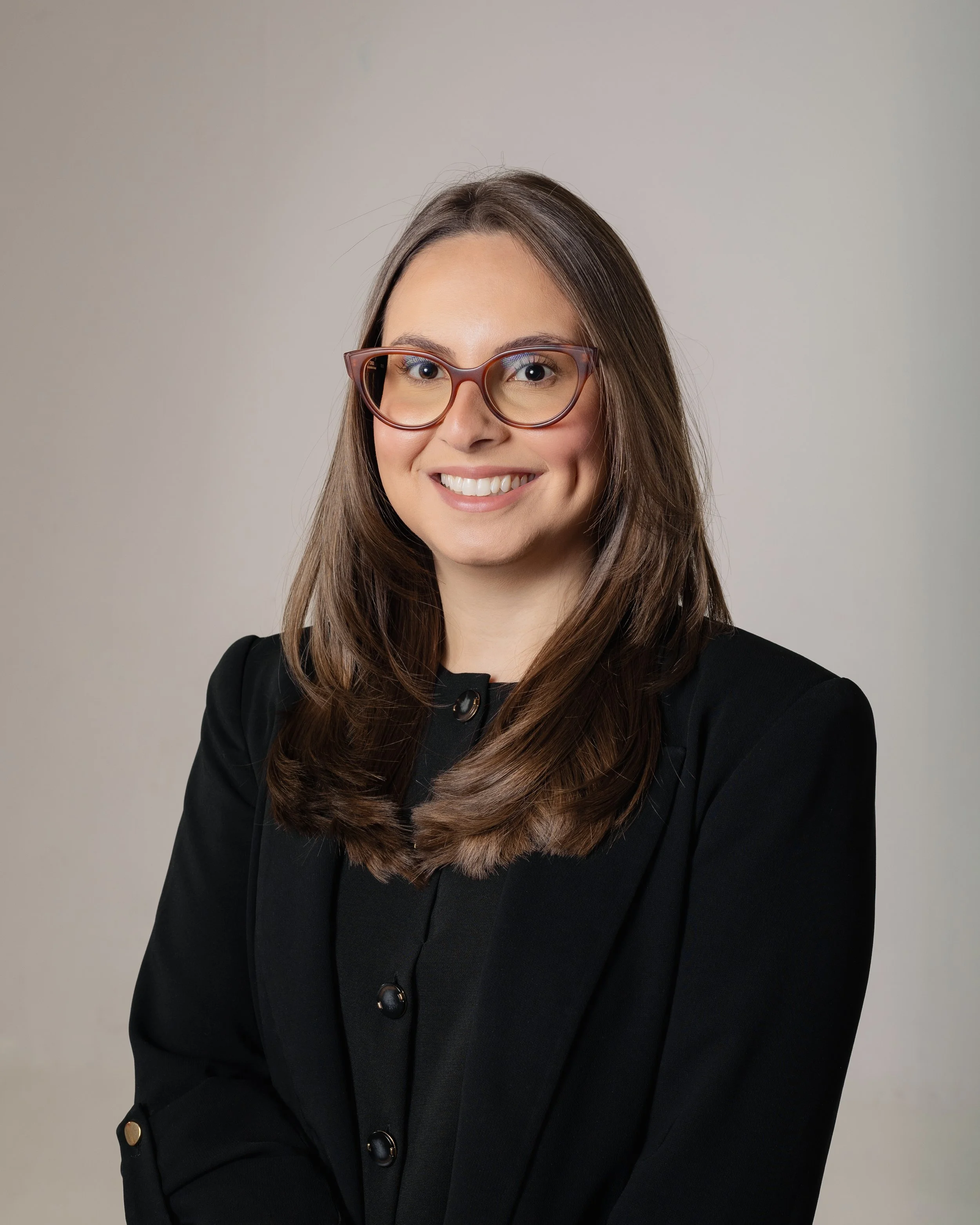 Homem sorridente de terno e gravata azul, com cabelo escuro e barba bem aparada, pose de perfil em fundo neutro.
