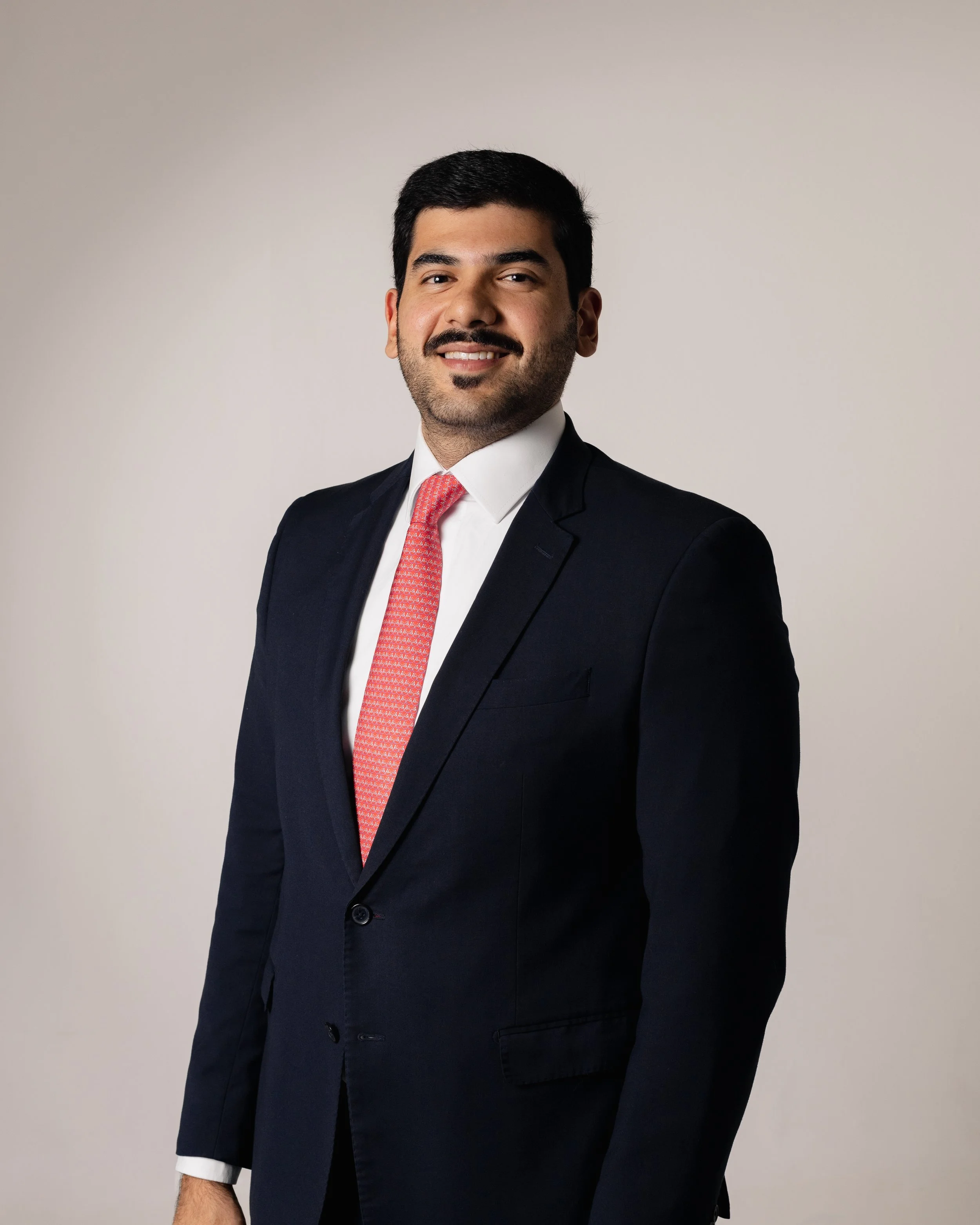 Homem sorridente de terno e gravata azul, com cabelo escuro e barba bem aparada, pose de perfil em fundo neutro.