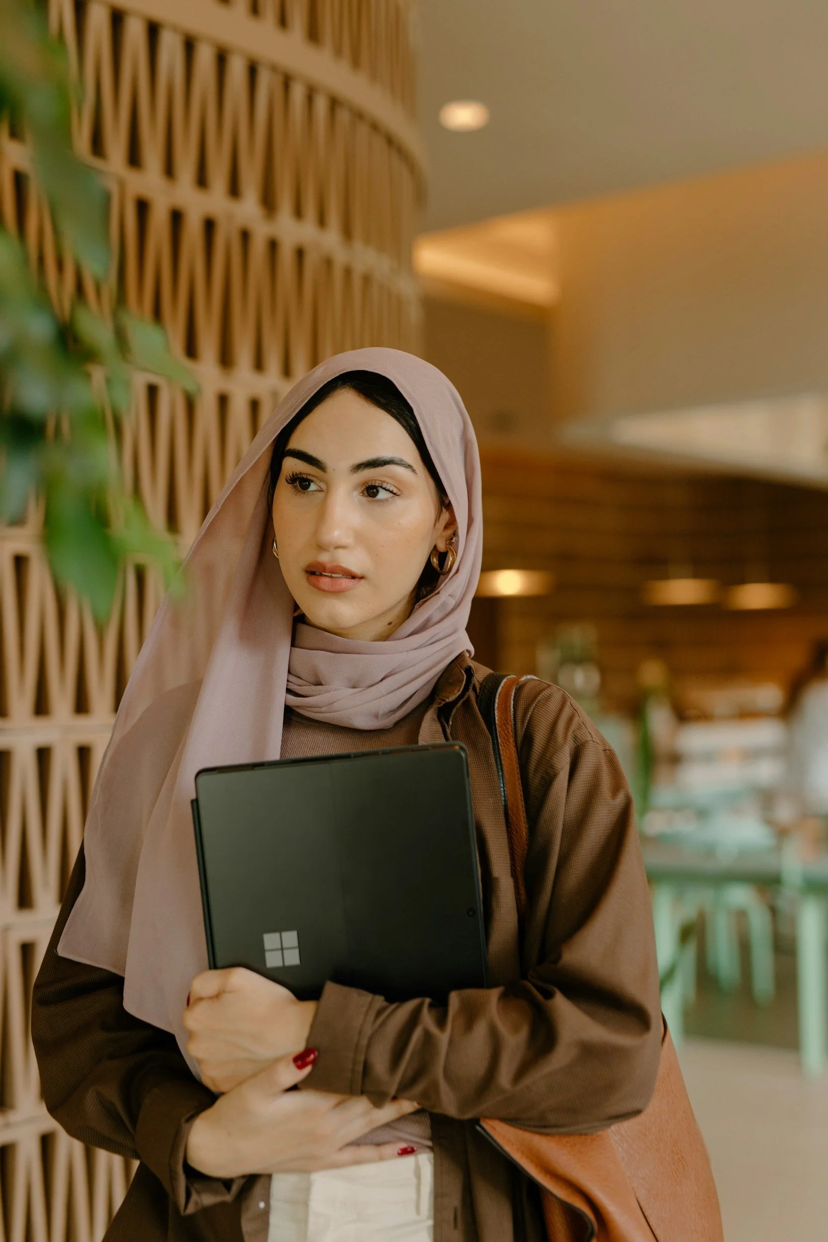 A young woman wearing a beige hijab and brown jacket holds a laptop close to her chest, standing in a warmly lit indoor space with wooden decor in the background.