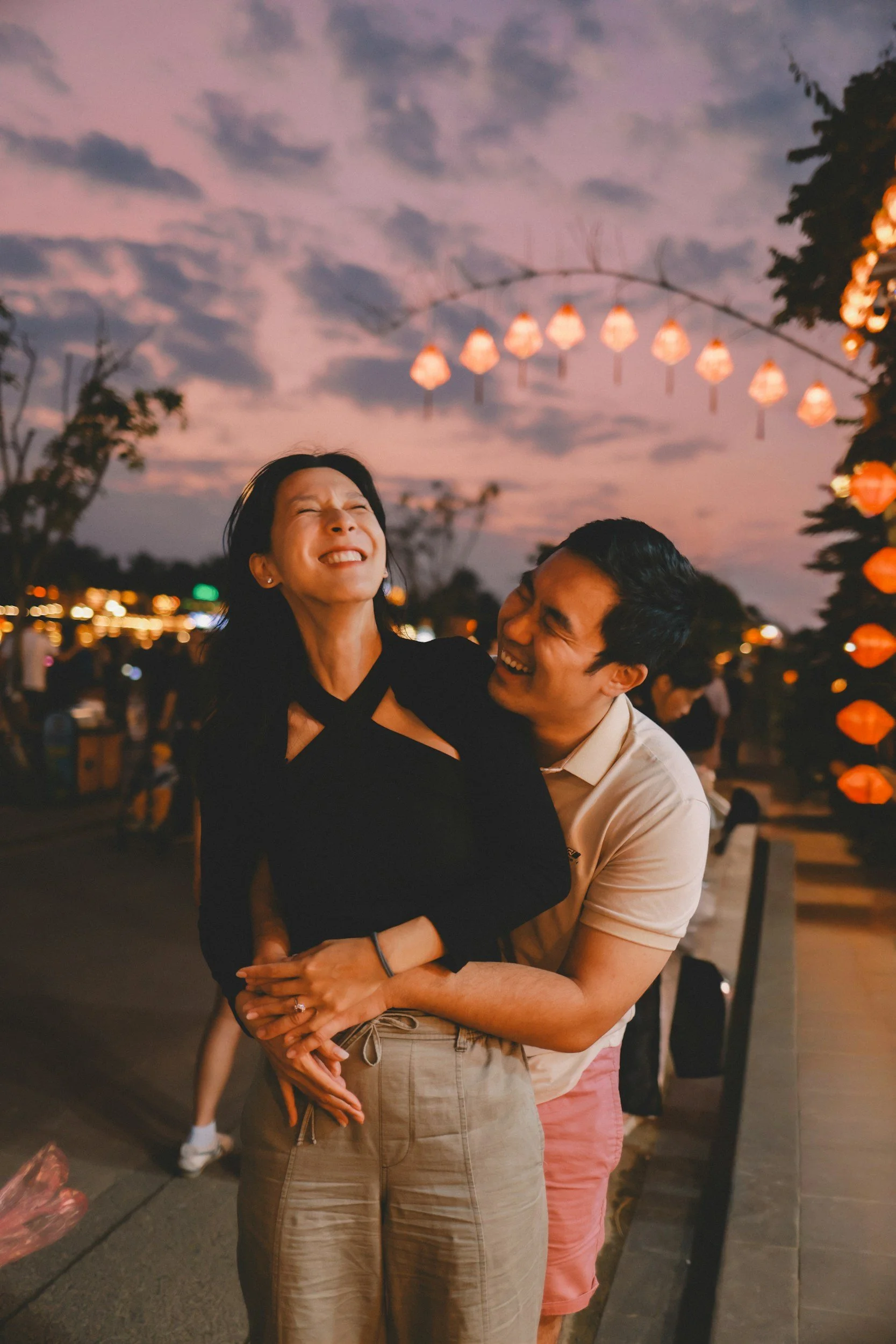 A woman and a man sharing a joyful moment at an outdoor festival during sunset, with hanging lanterns and other people in the background.