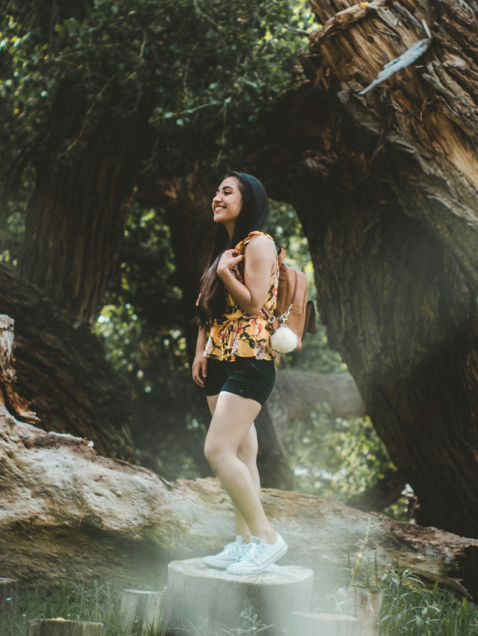 A young woman with long dark hair and tan skin standing on a tree stump outdoors, smiling and carrying a backpack, surrounded by trees and greenery.
