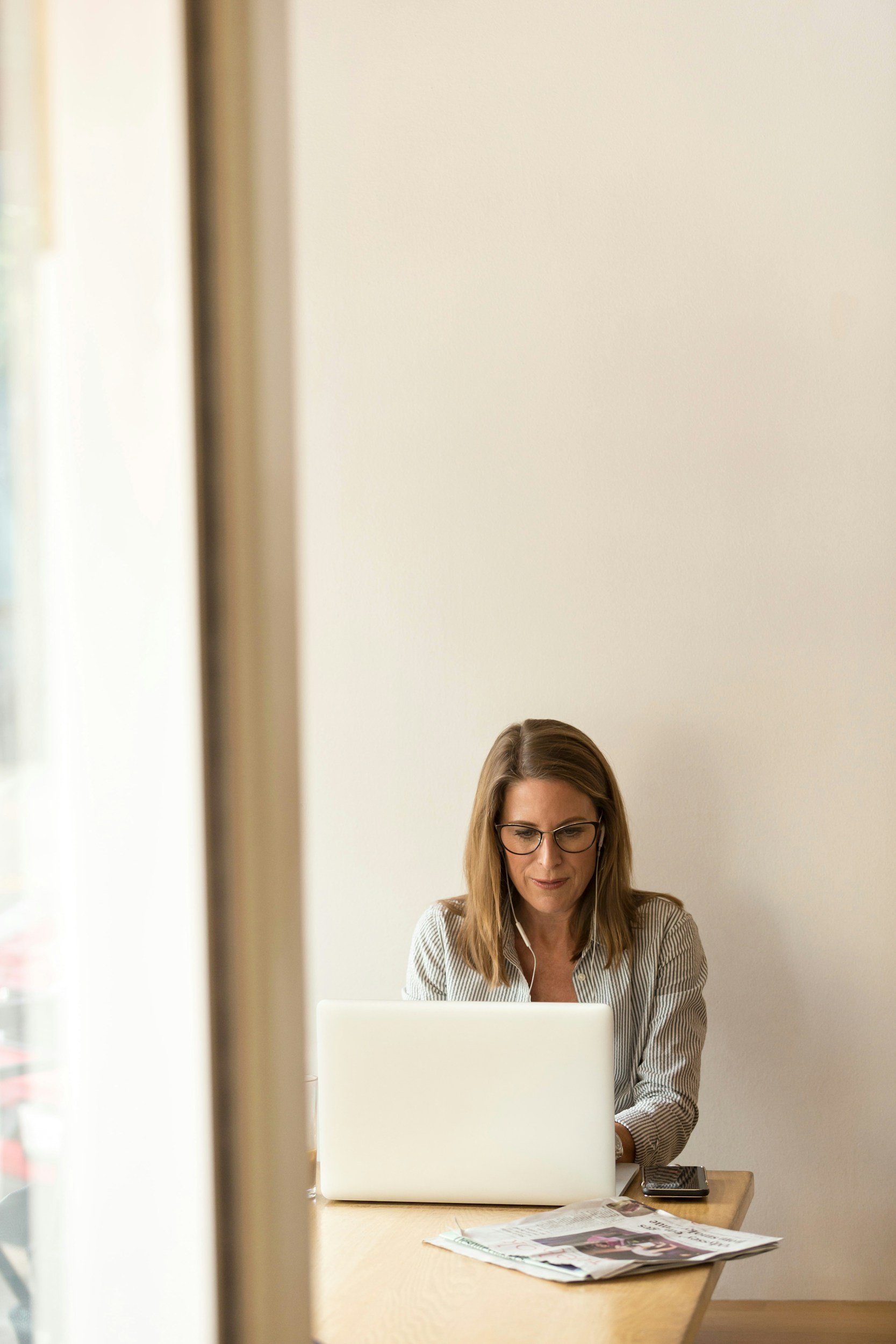 A woman with glasses working on a laptop at a desk, with newspapers and a phone in front of her, in a brightly lit room.