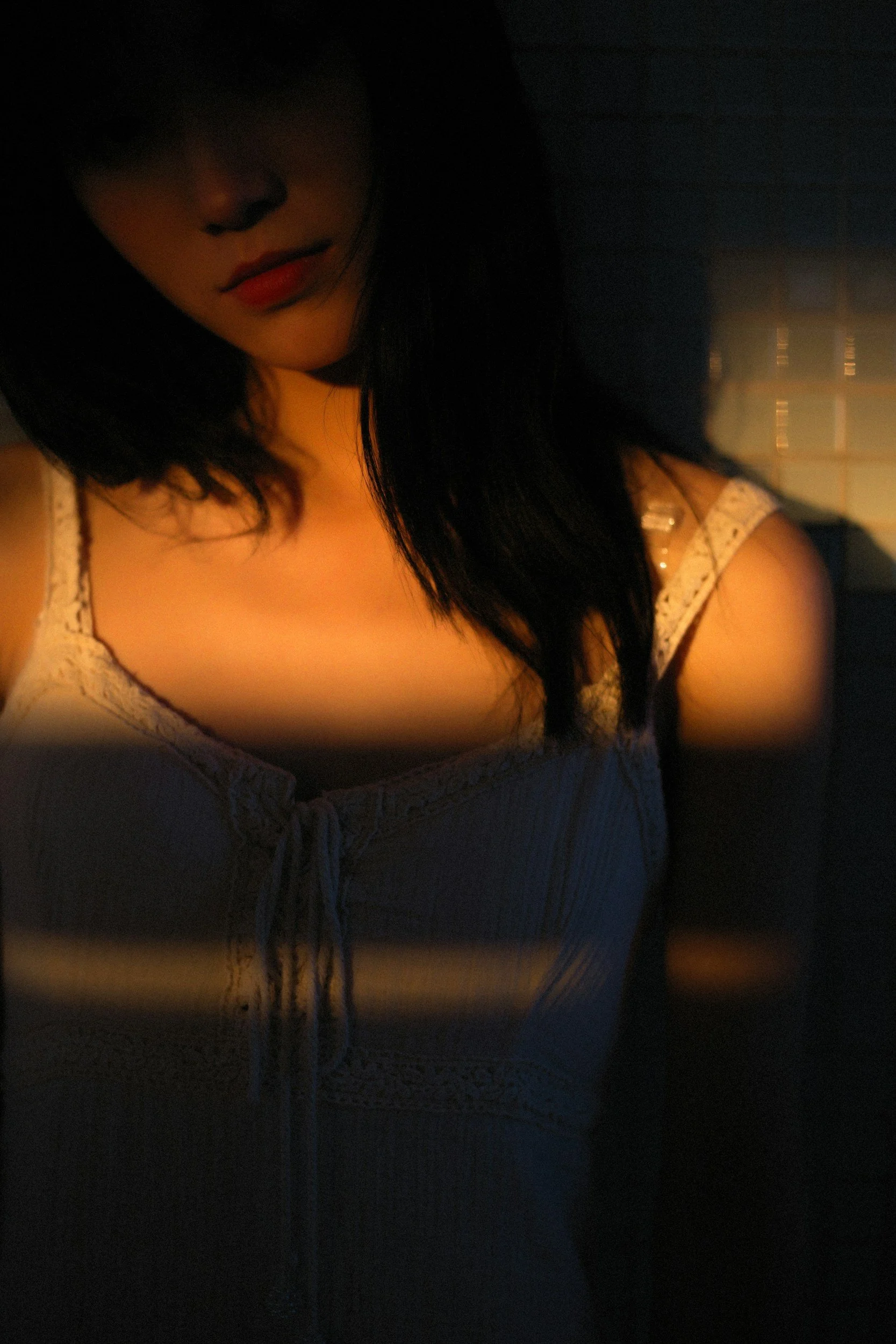 A woman with dark hair and red lipstick wearing a light-colored, lace-trimmed top, standing in a dark room with sunlight filtering through blinds.