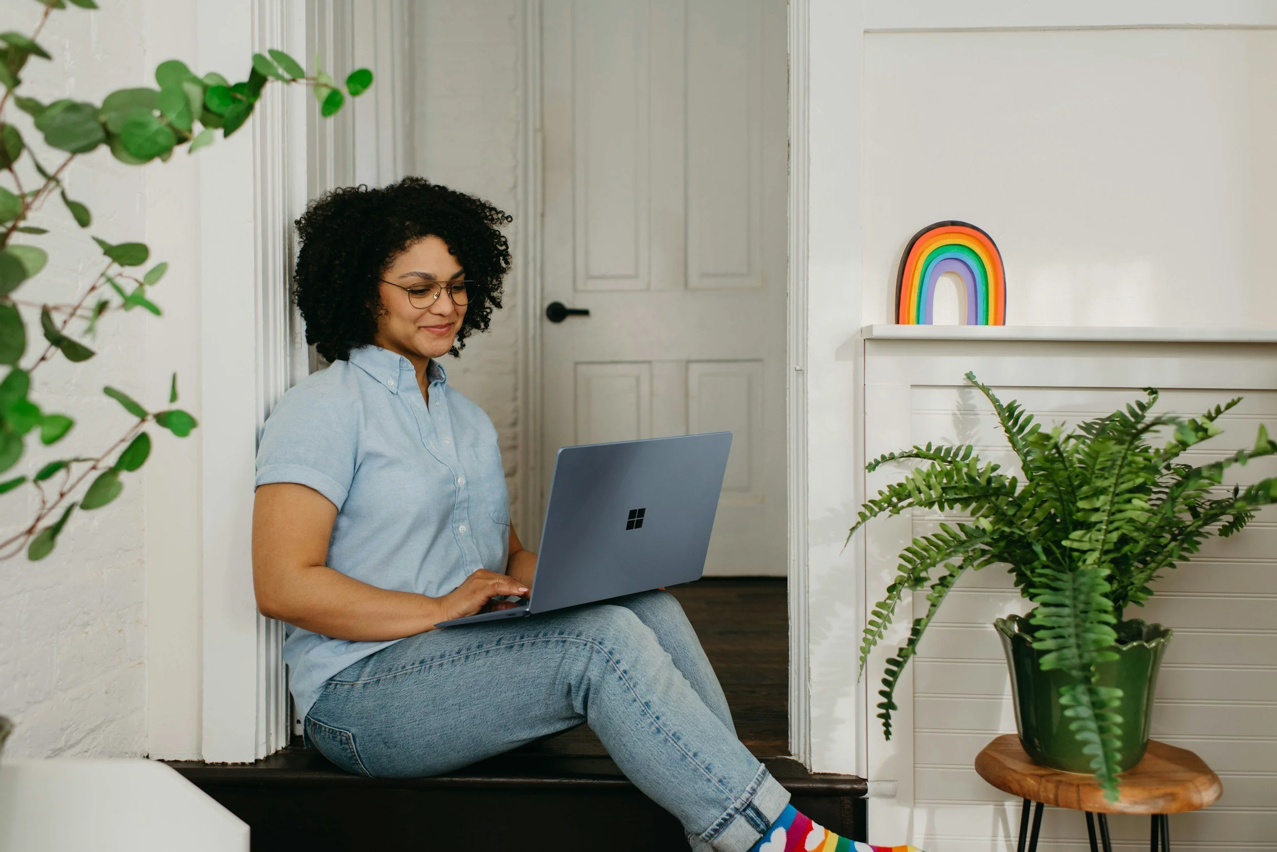 A woman with curly hair and glasses sits on a staircase, working on a silver laptop. She is wearing a light blue shirt and jeans. There are houseplants nearby, and a colorful rainbow decoration on the wall behind her.