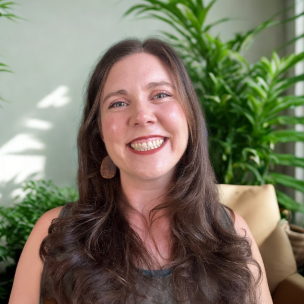 A woman with long dark hair smiling in a room with green plants and a beige chair.