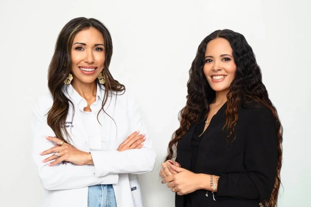 Two women smiling, one in a white professional blazer with crossed arms, the other in a black blazer with hands clasped, against a plain white background.