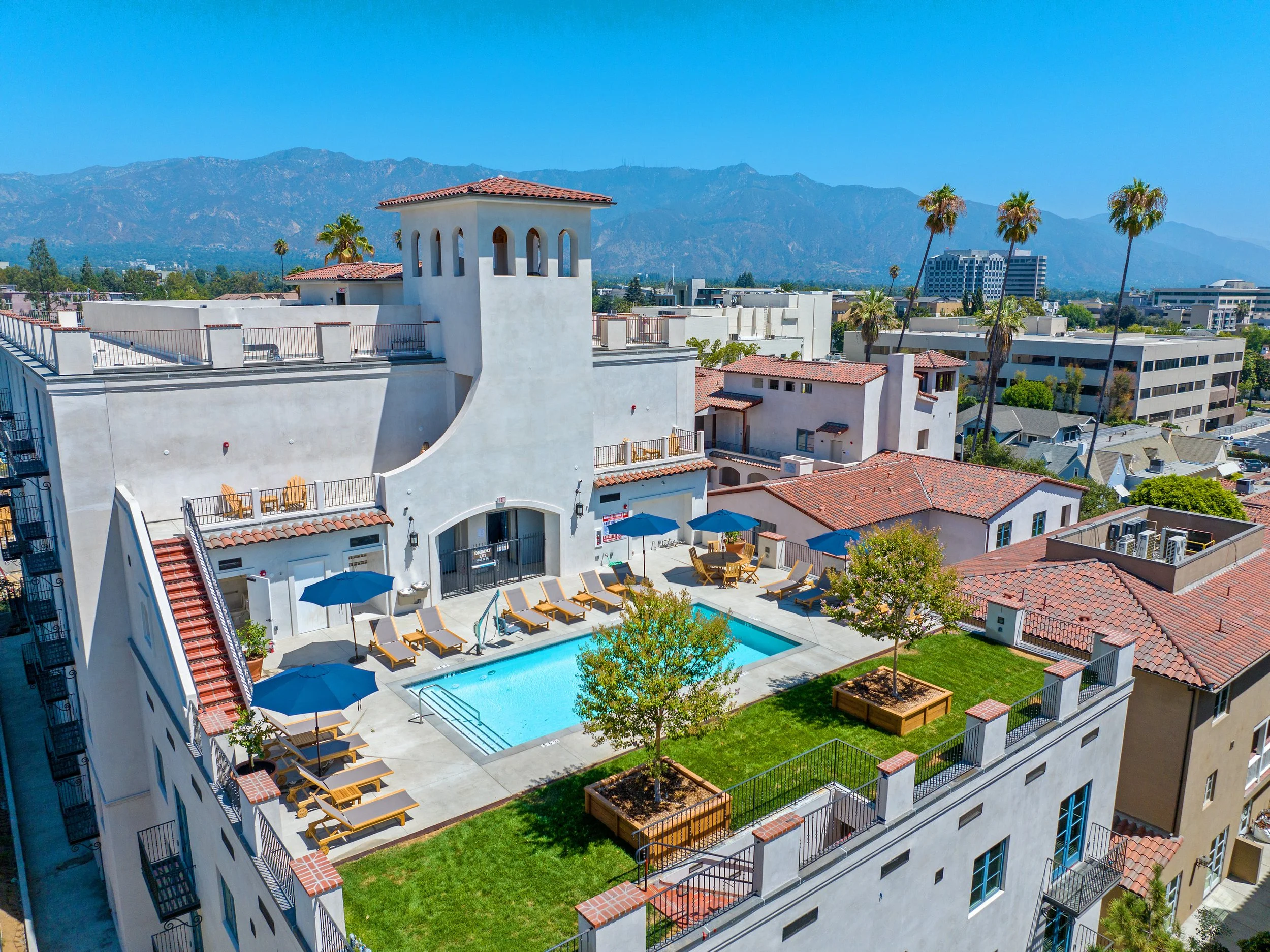 Aerial view of Catalonia rooftop pool and landscaped terrace with lounge seating, umbrellas, Spanish-style architecture, and mountain views.