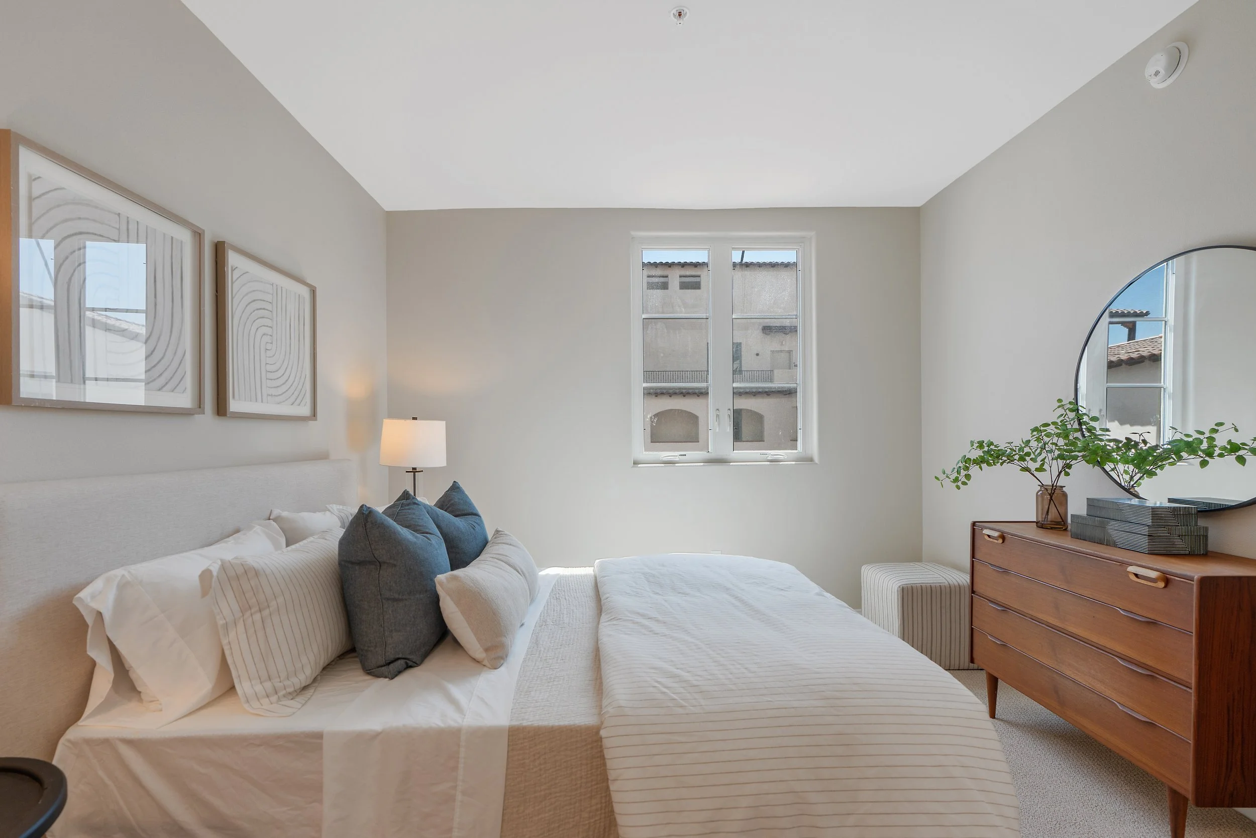Bright bedroom at Catalonia featuring a neutral upholstered bed, wood dresser, round mirror, framed wall art, and natural light.