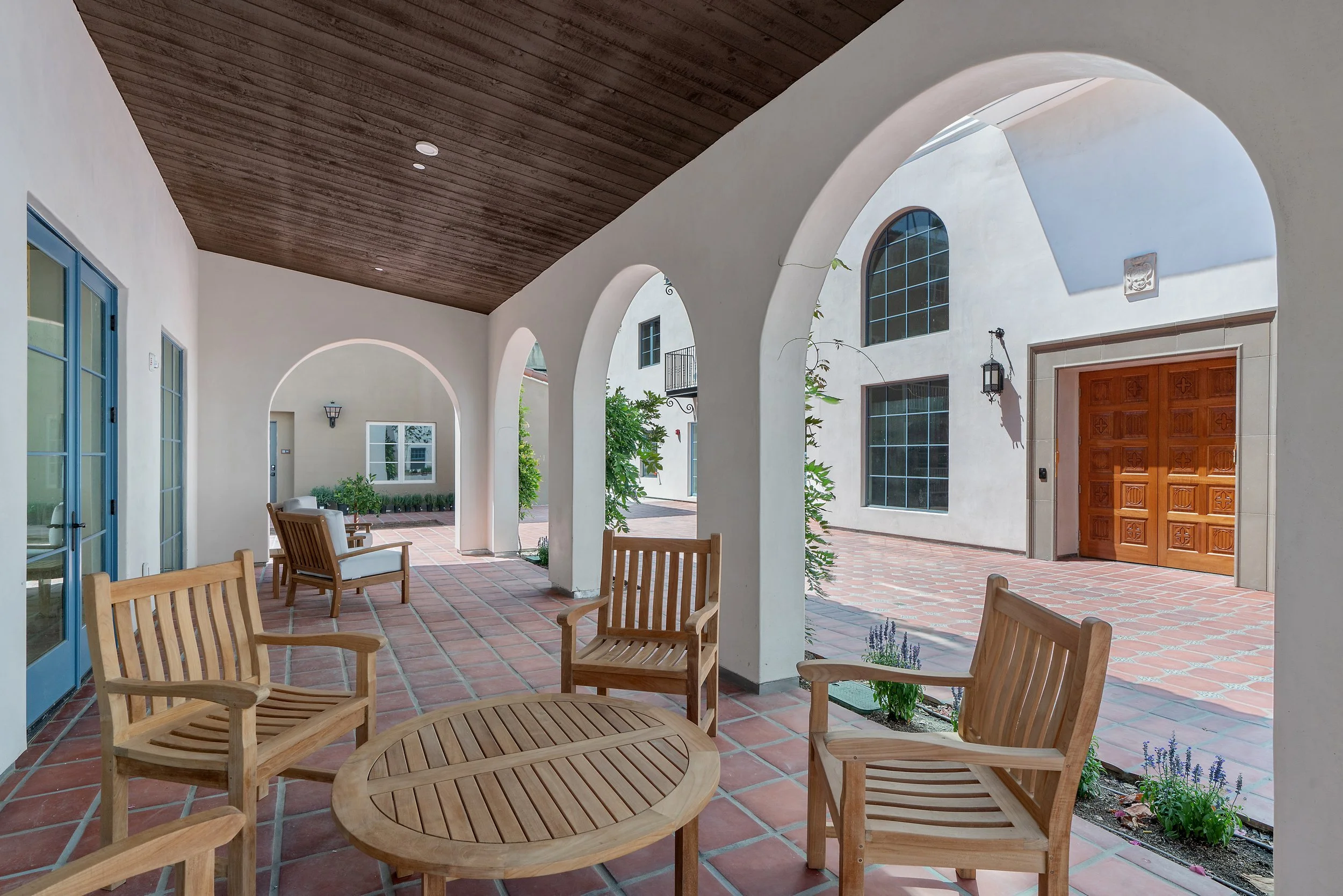 Covered courtyard seating area at Catalonia Apartments in Pasadena, CA, featuring Spanish-style arches, terracotta tile flooring, and wooden lounge chairs.
