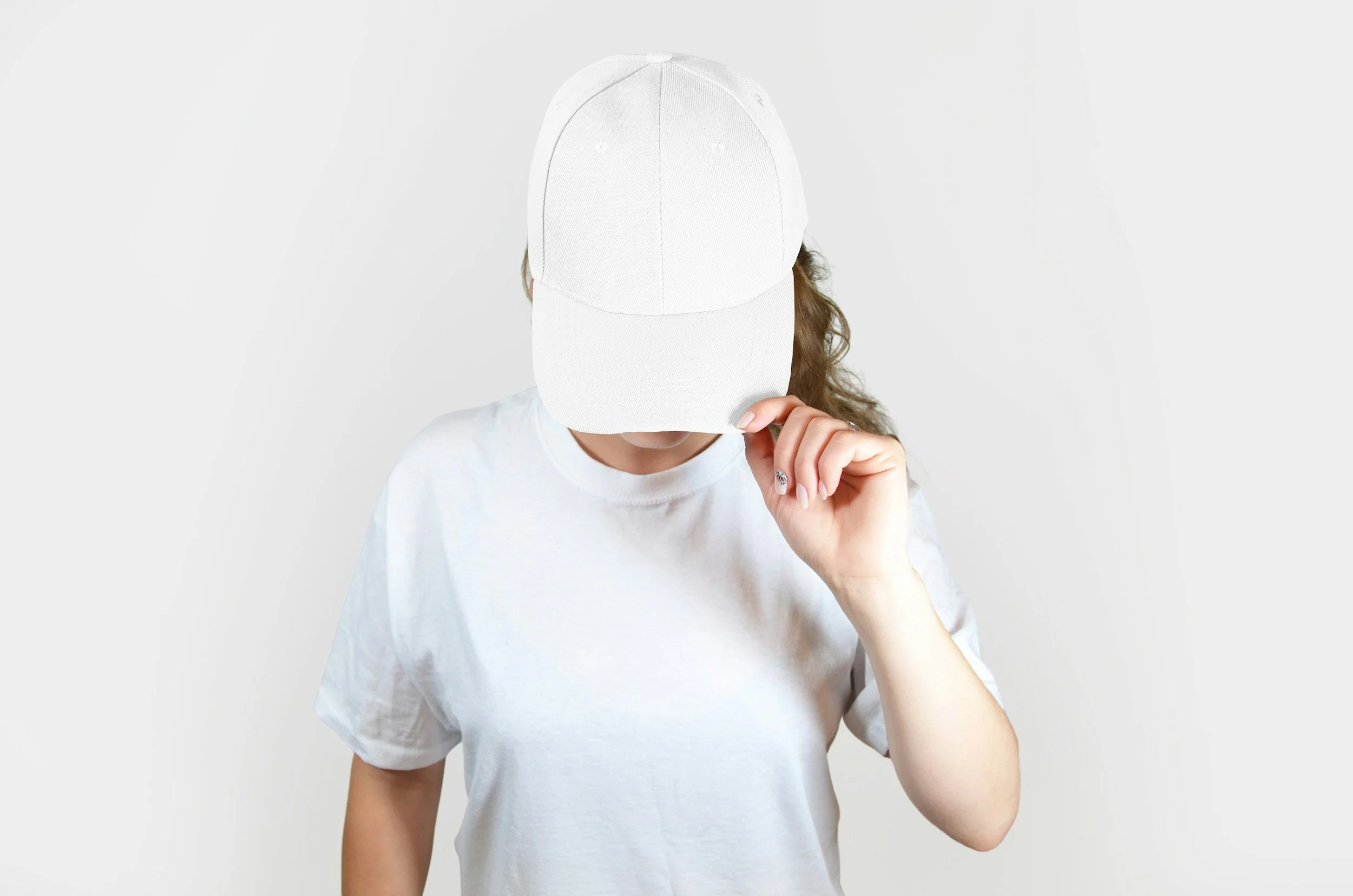 Person wearing a white baseball cap and white t-shirt, with long curly hair, standing in front of a plain white background.