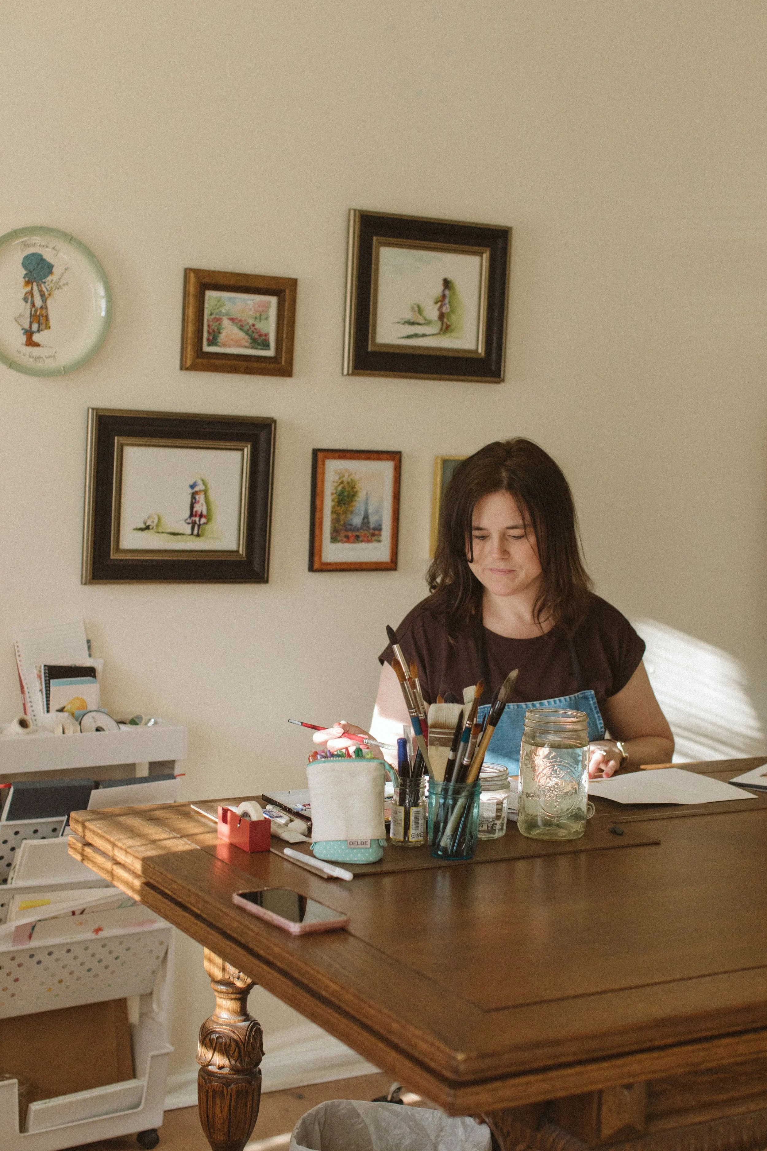 A woman with dark hair and a brown shirt sitting at a wooden table surrounded by art supplies, looking at her phone, with a decorated wall behind her.