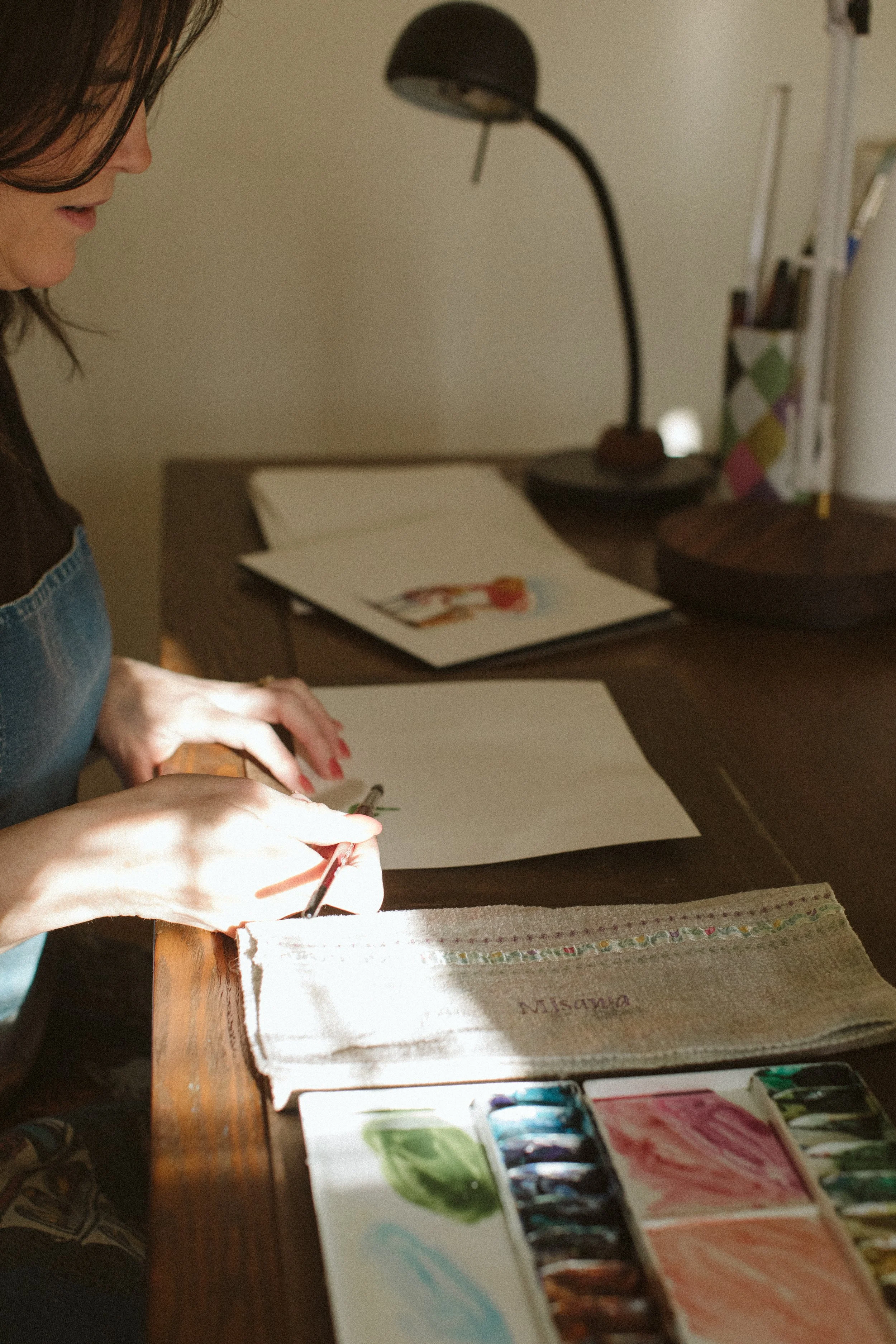 Keri Misawa, watercolor artist in Omaha, NE, paints at a wooden table, with painting supplies