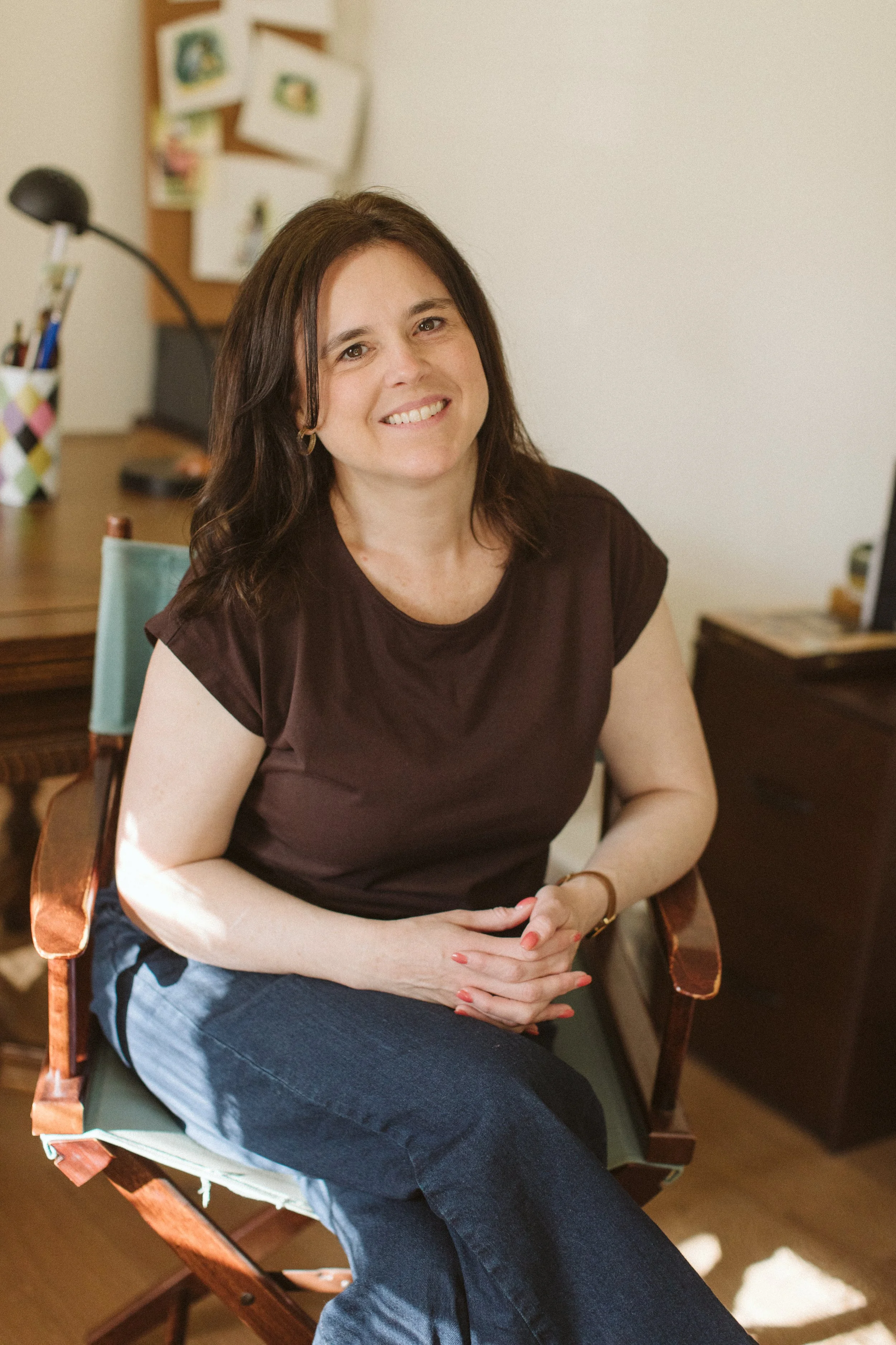 A woman with shoulder-length dark hair smiling, sitting on a wooden chair, wearing a dark brown top and blue jeans, in a room with a desk and art board in the background.
