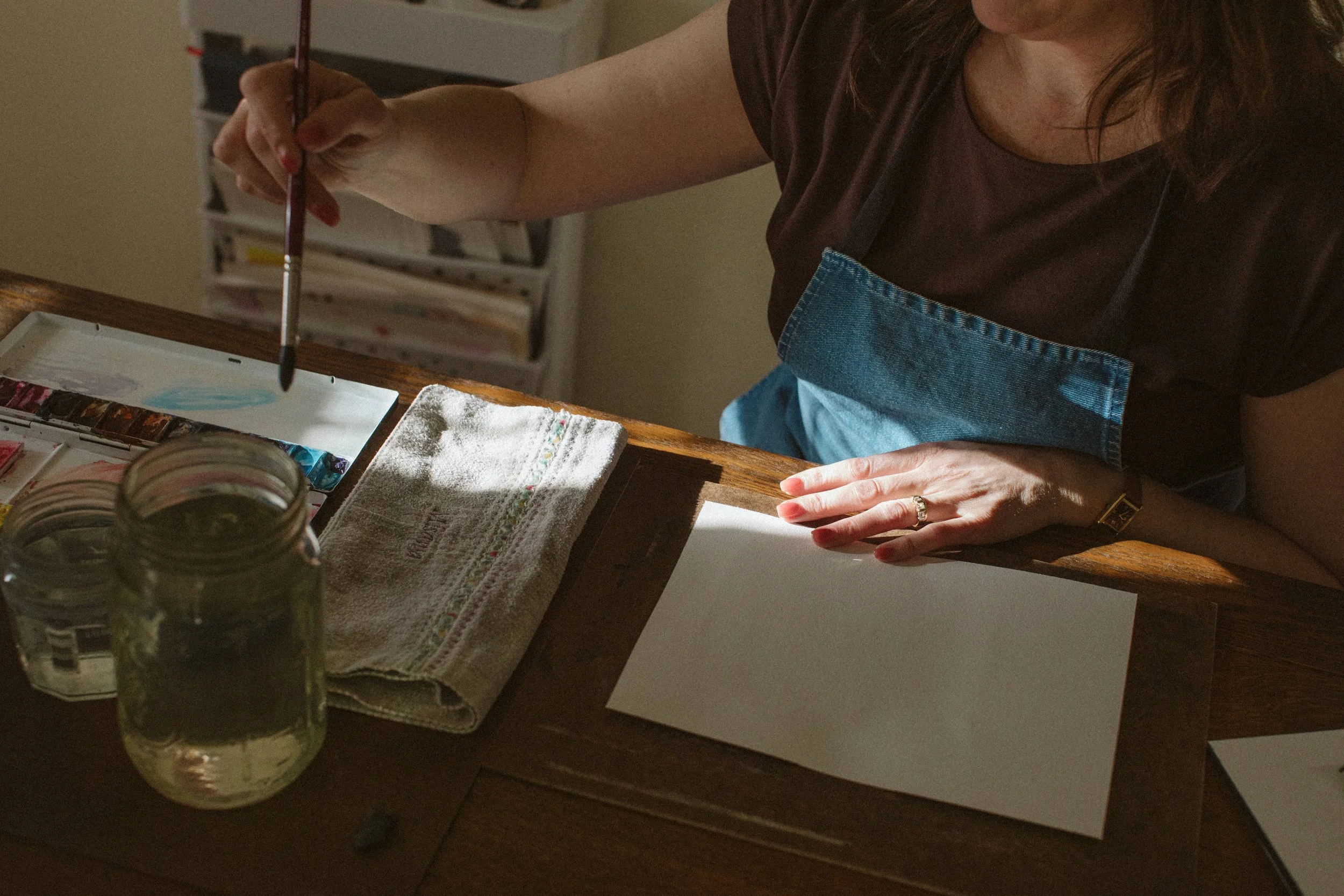 Person painting on a blank canvas at a wooden table with art supplies, jars, and a towel nearby.