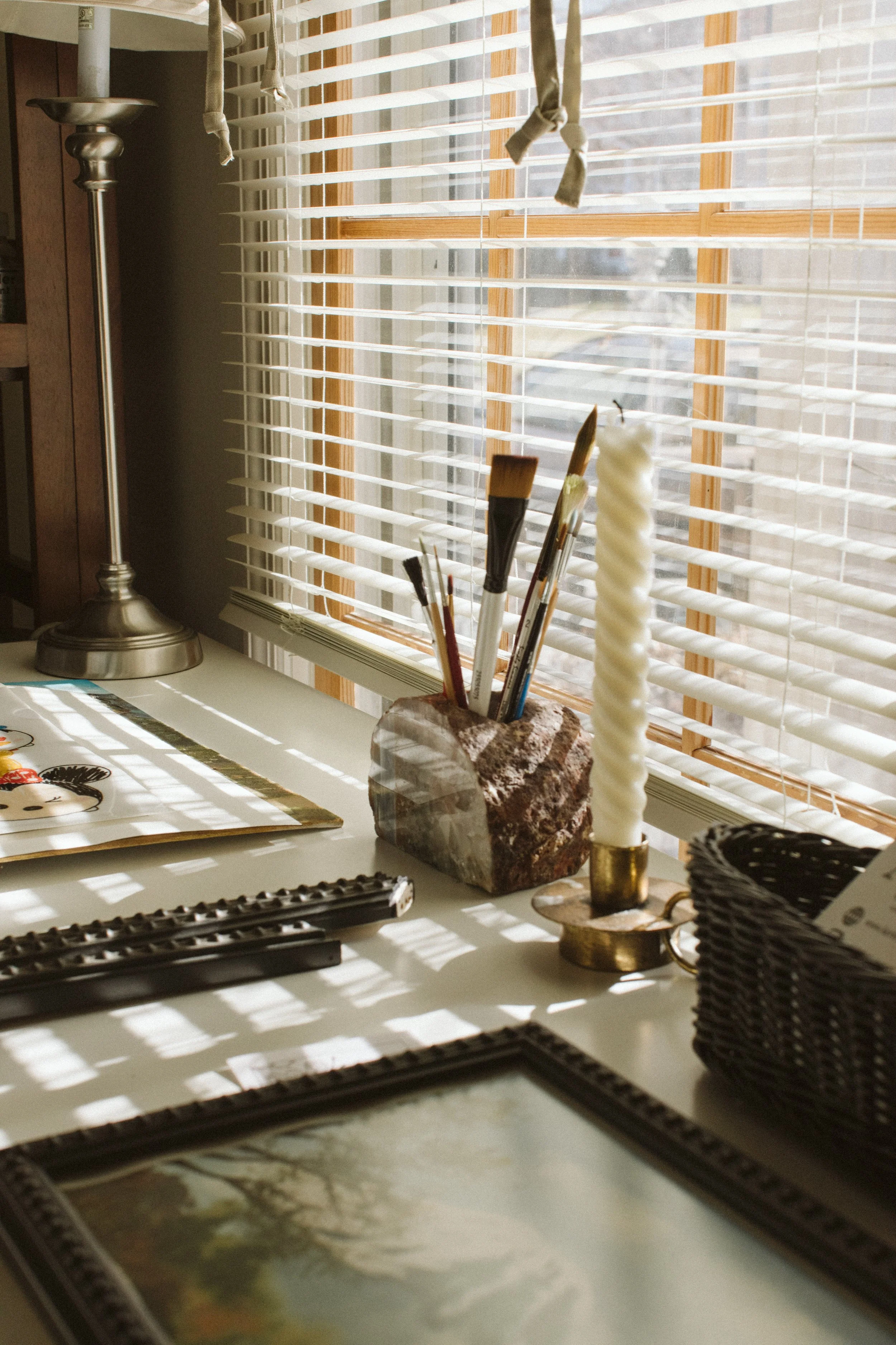 A window with wooden blinds casting shadows on a white desk. On the desk, there are paintbrushes in a stone holder, a brass candlestick, a magazine, and a woven basket.