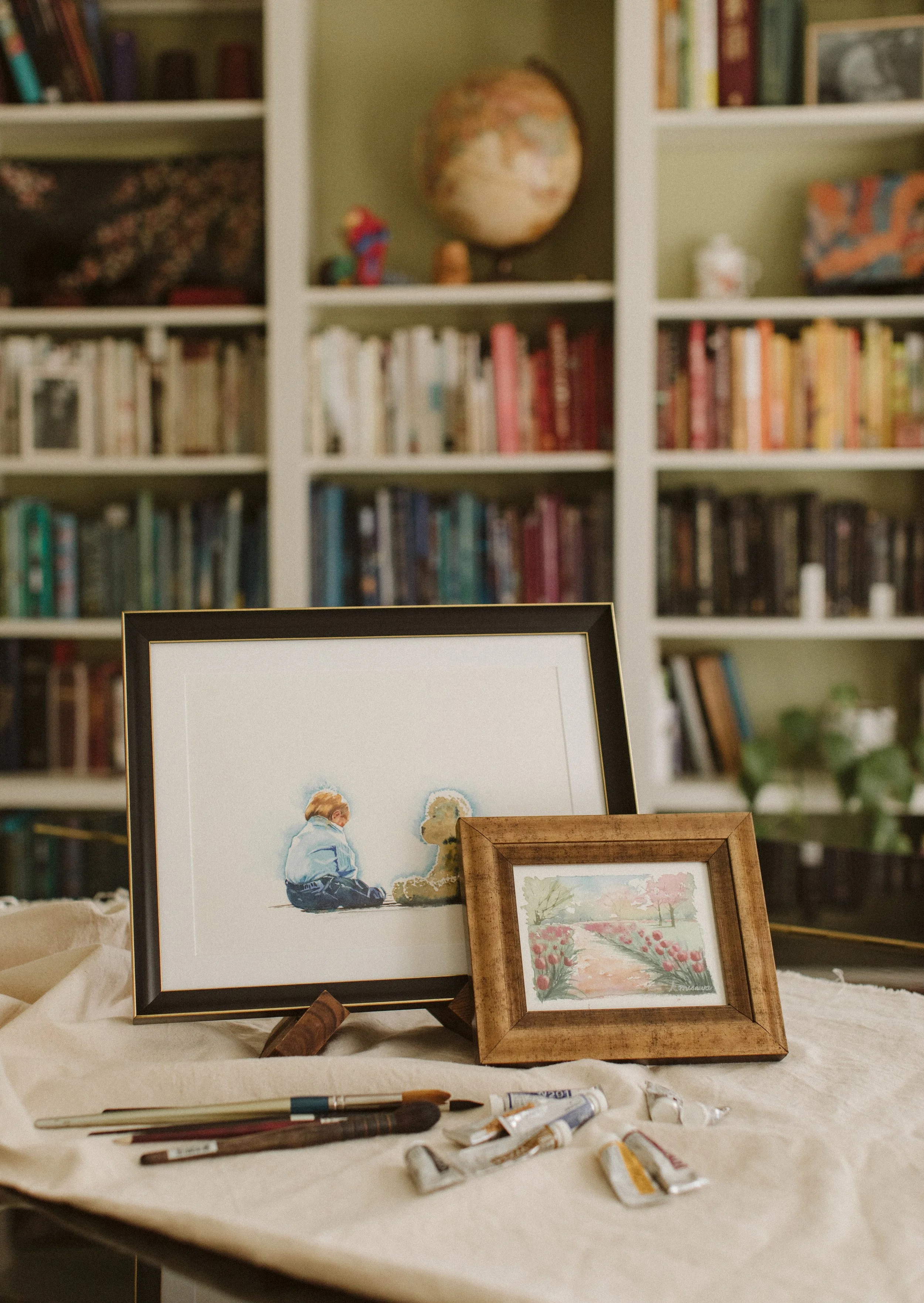 Painting supplies including brushes and tubes of paint on a table, with framed photographs and artwork in the background.