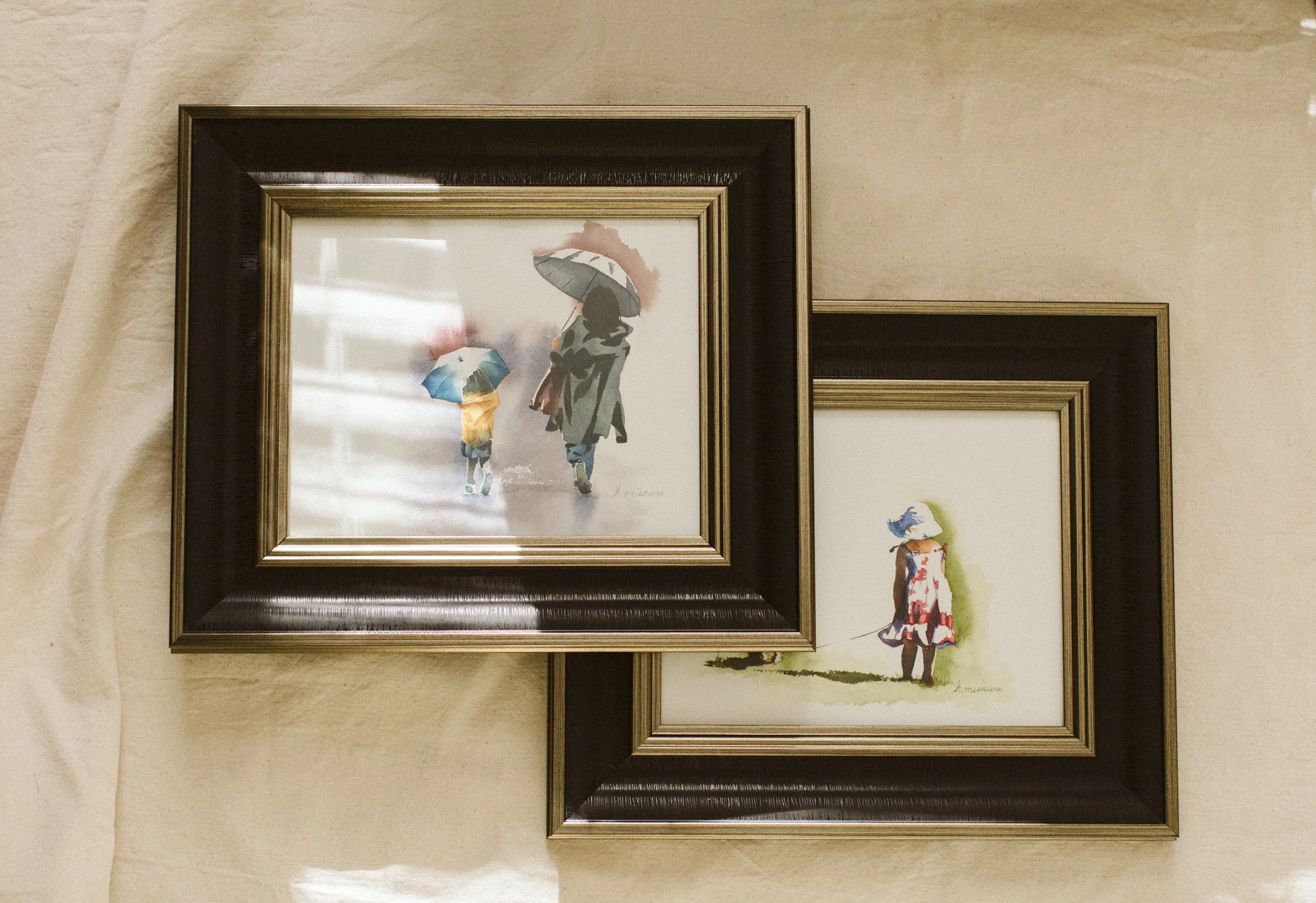 Two framed watercolor paintings of a child and mother holding umbrellas and a child in a wearing a dress and sun hat and holding a puppy on a leash painted by Keri Misawa, watercolor artist in Omaha, NE