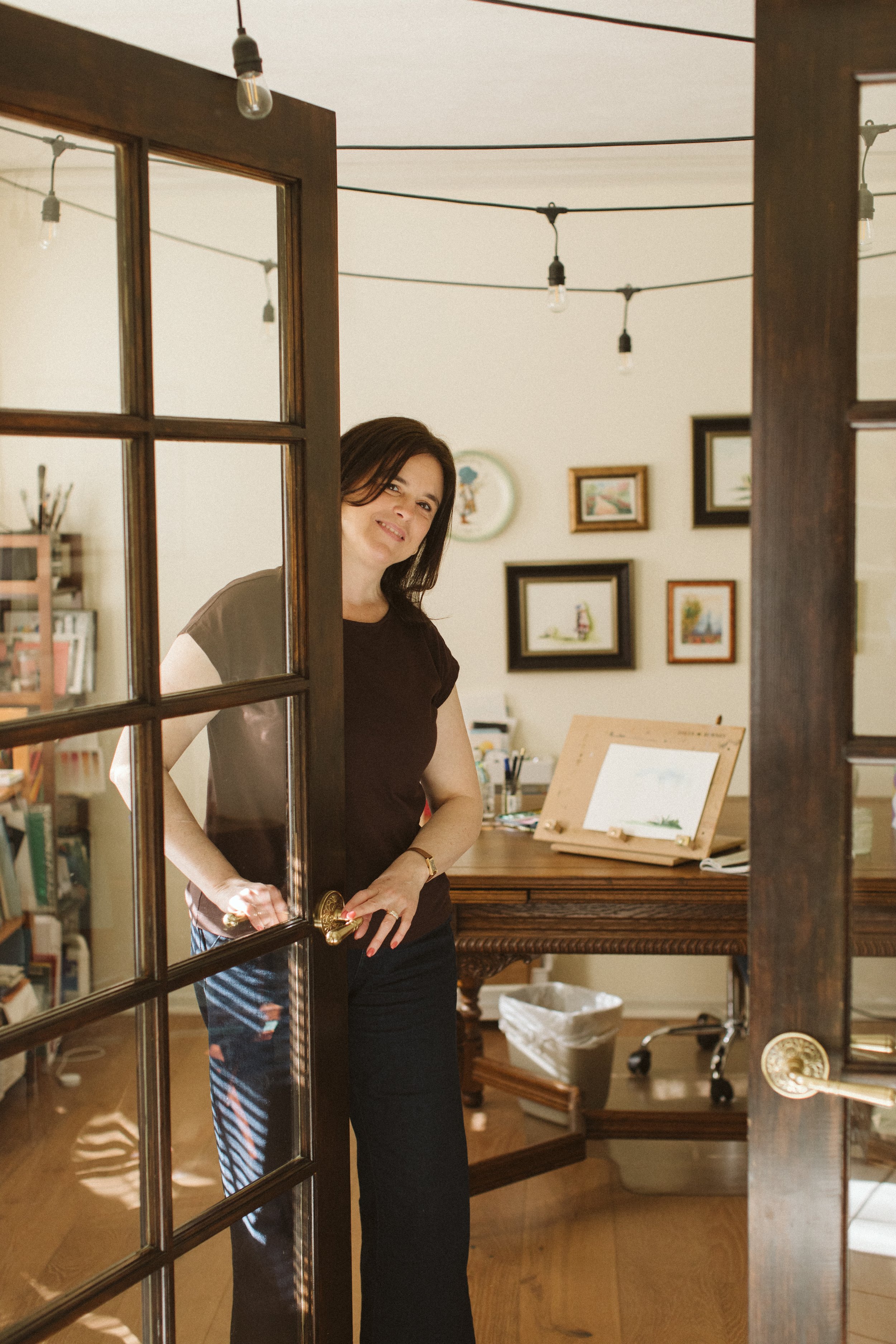 Keri Misawa, watercolor artist in Omaha, NE, standing in her sun-lit studio with her desk of art supplies and her original paintings and other works of art on the wall behind her.