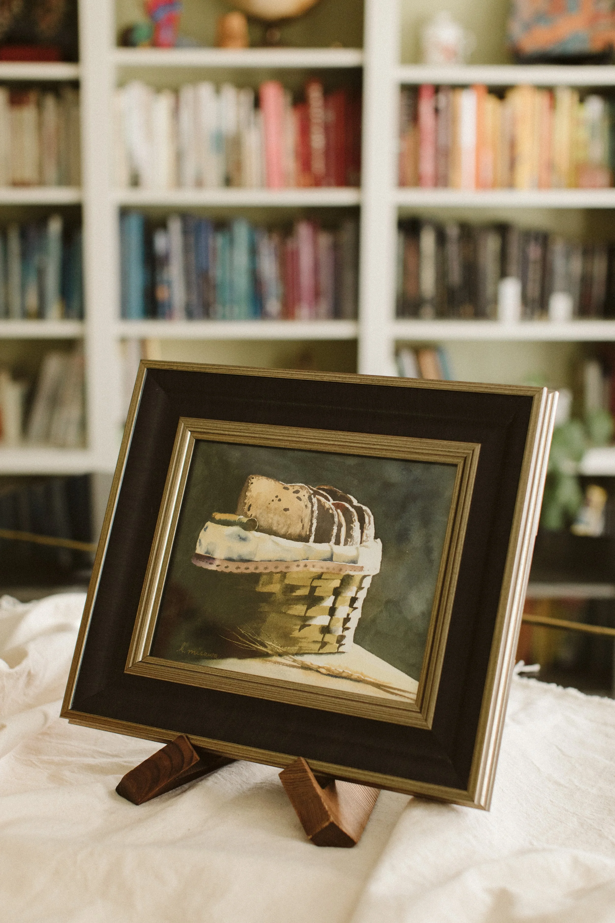 Framed watercolor painting by Keri Misawa, watercolor artist in Omaha, NE of a basket full of sliced bread on a wooden easel, with a bookshelf filled with books in the background.