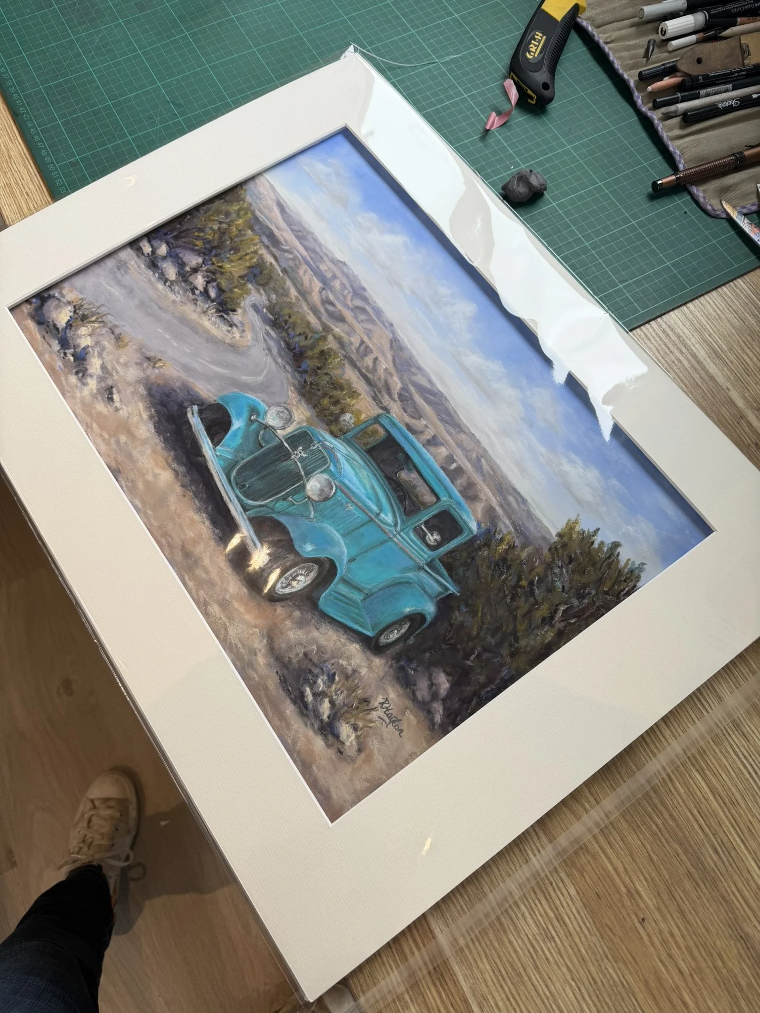 Artist's workspace with a framed painting of a turquoise vintage race car on a mountain road, surrounded by drawing tools and a cutting mat.