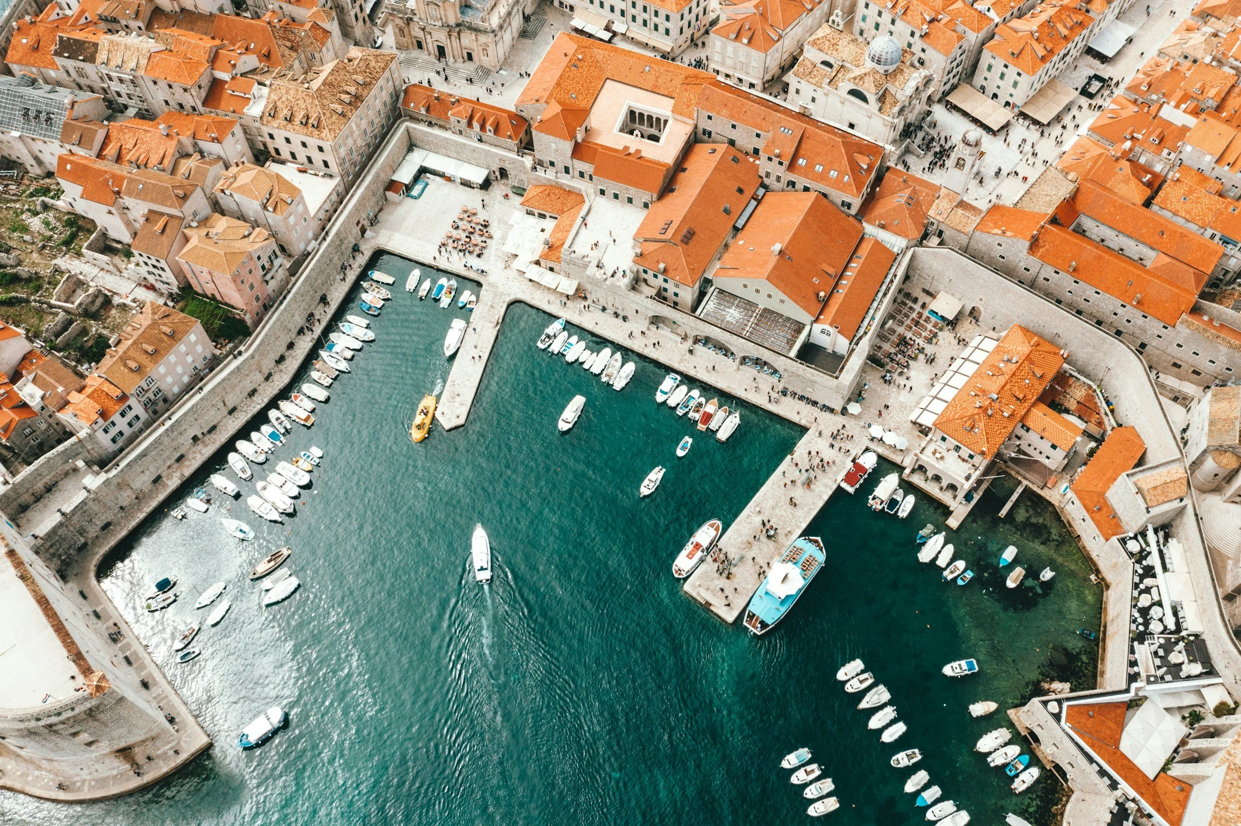 Aerial view of a coastal city with red-tiled roofs, enclosed harbor with boats, and stone walls along the waterfront.