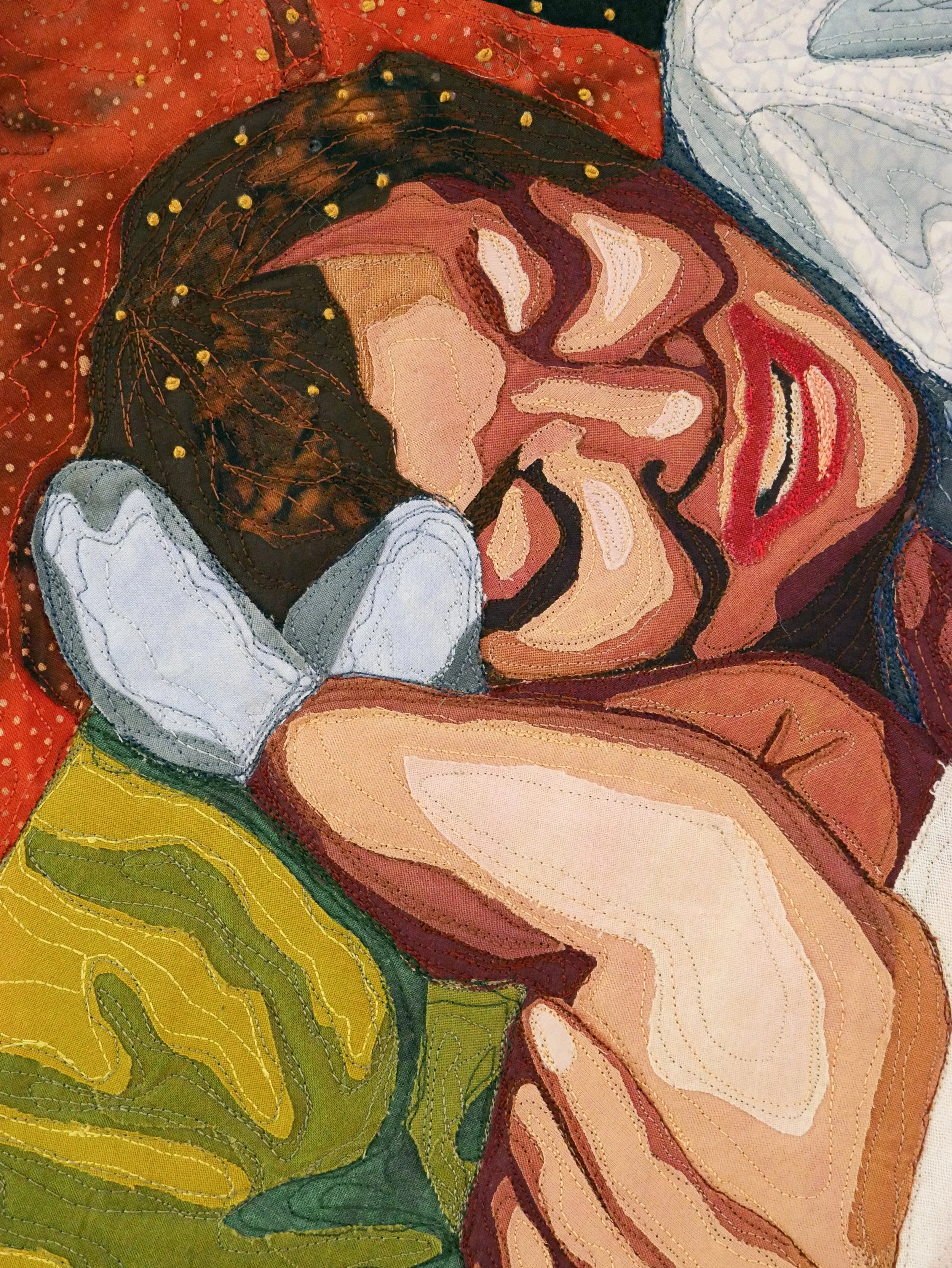 Quilt detail of the grandma's face resting on an orange pillow next to her infant grandchild's legs