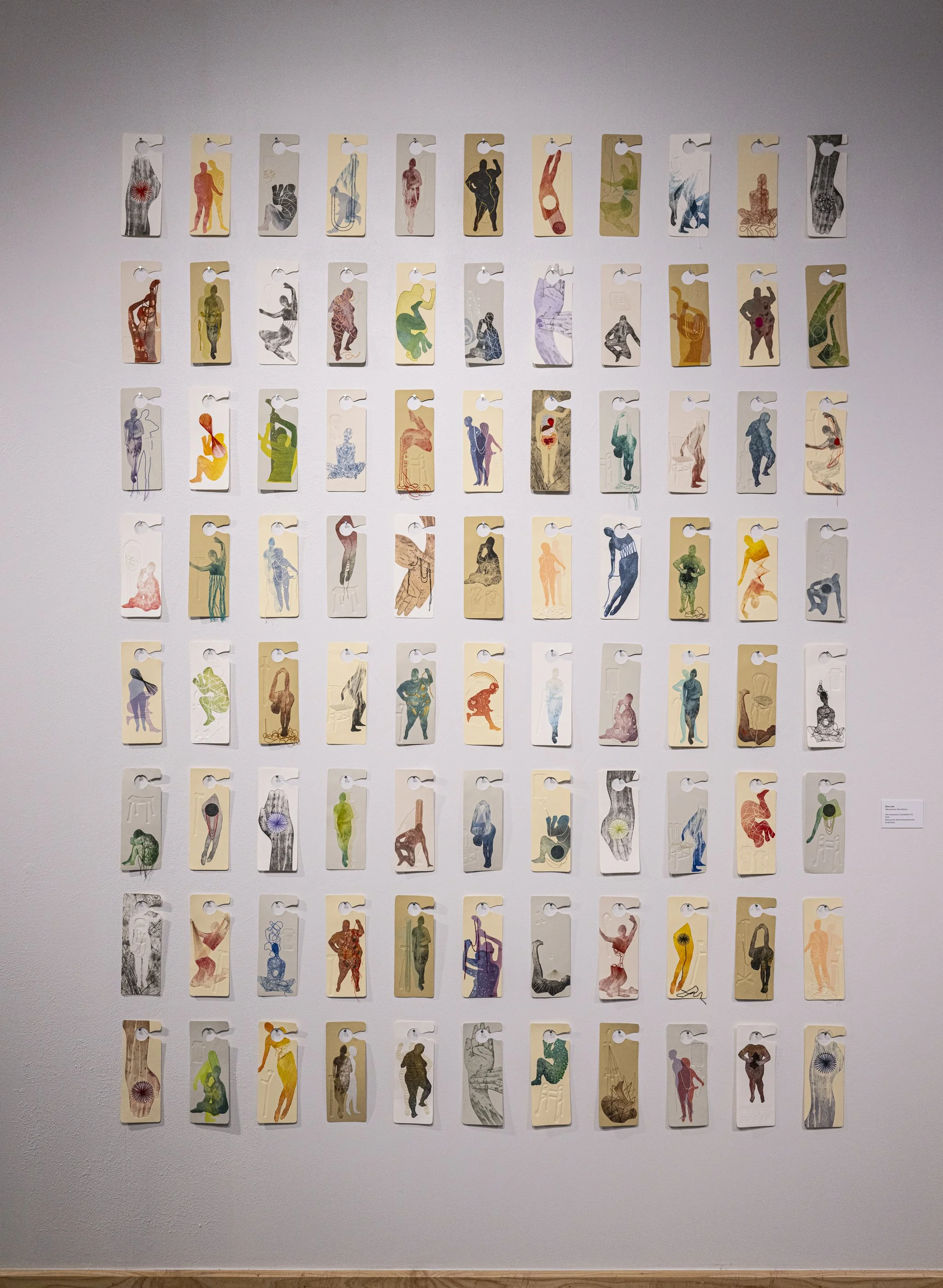 A print installation comprised of a grid of multicolored prints shaped like accessible parking placards. Each print is unique and features a disabled figure in different positions, some with embossed mobility aids and colorful embroidery.