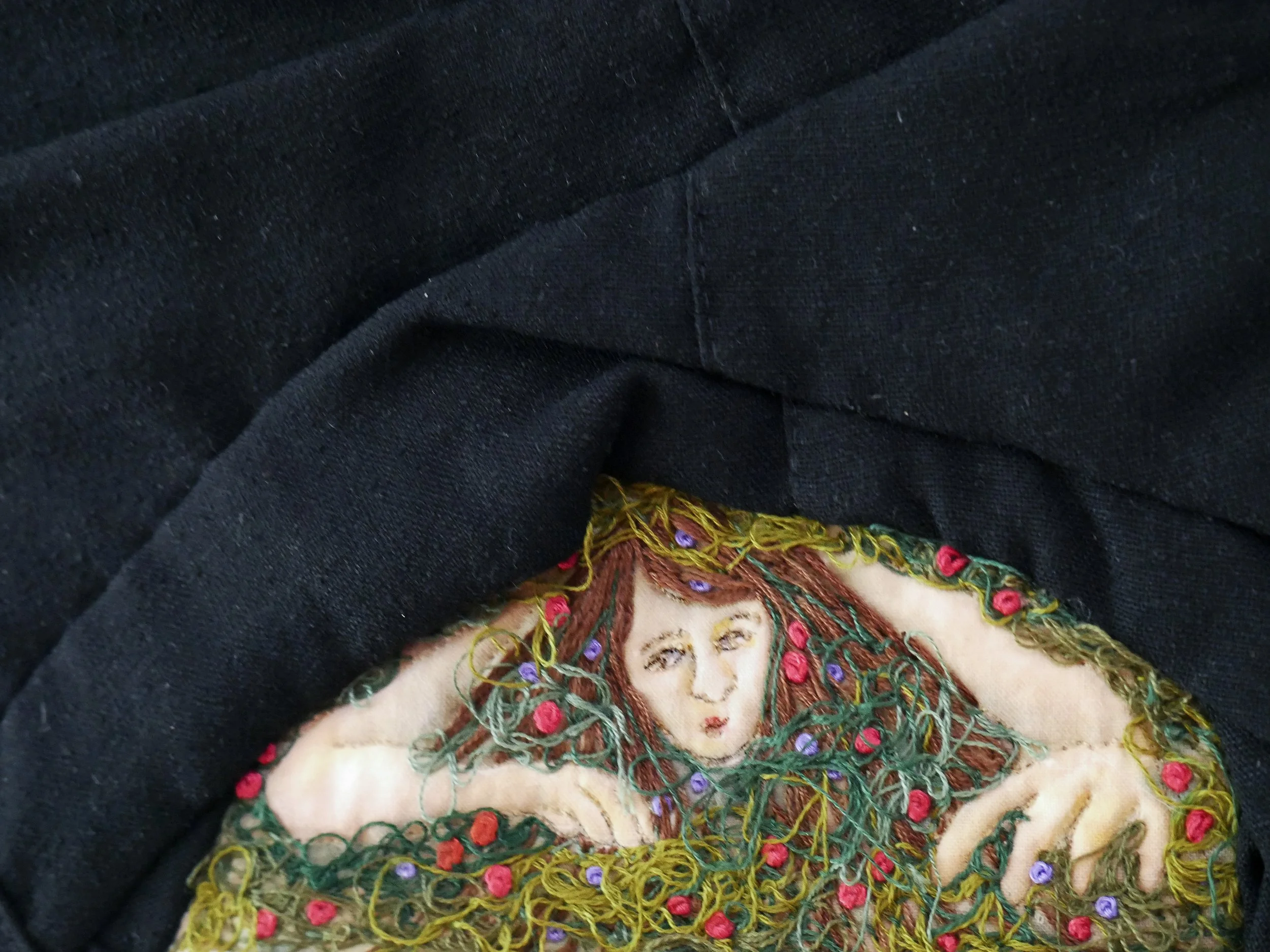 Detail of a figure with brown hair crawling out from under black fabric and surrounded in moss and flowers