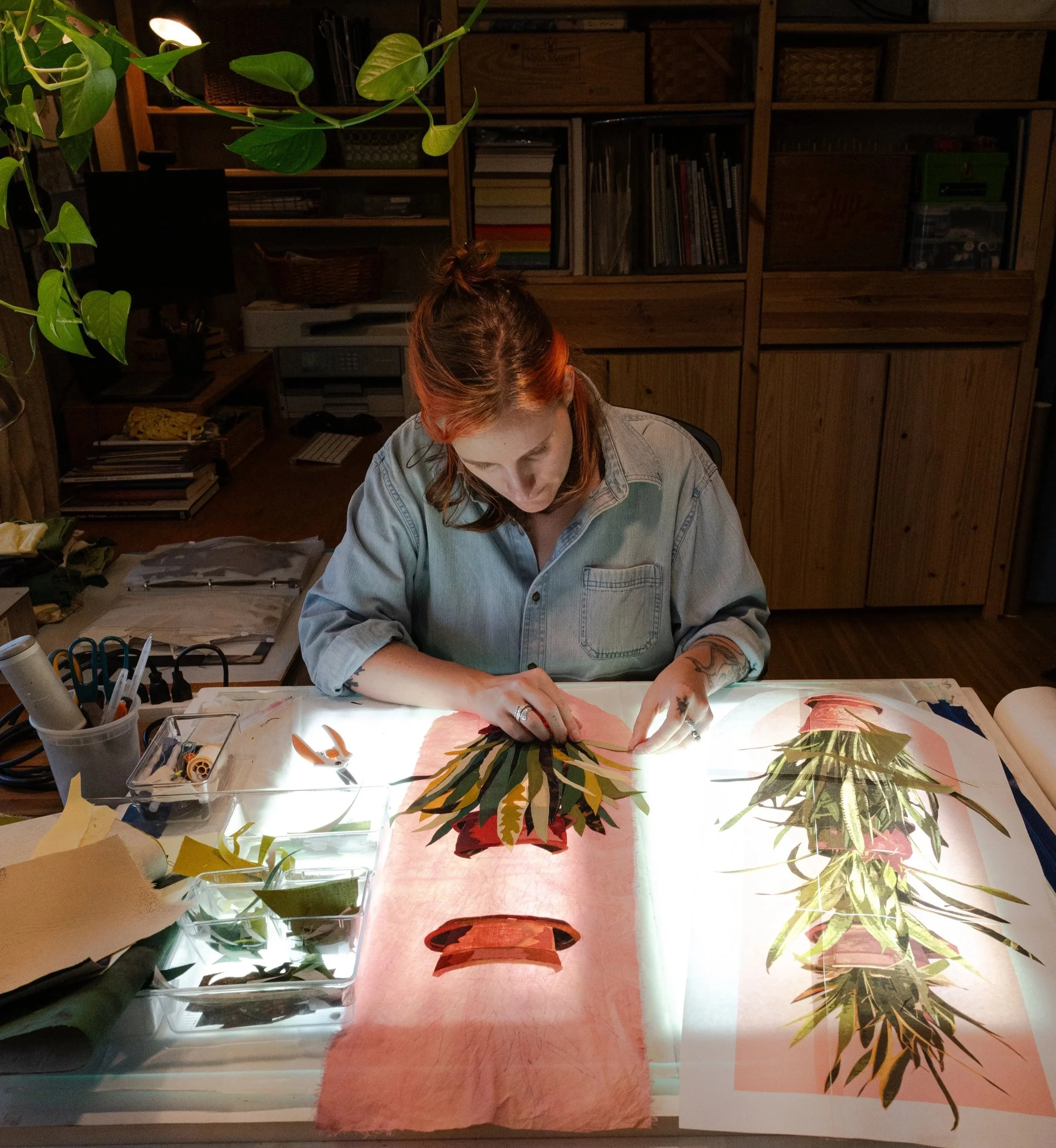 Eliza, a white femme person with orange hair in a denim button up, leans over a light table as they work on a fabric collage of a snake plant on a peach backing fabric.