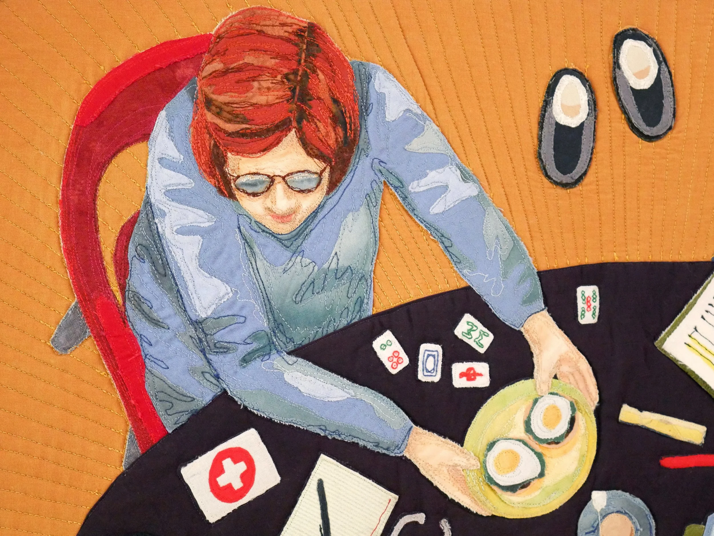 Quilt detail of a woman with red hair in a blue shirt sitting in a red chair at a dinner table holding a plate of fried eggs