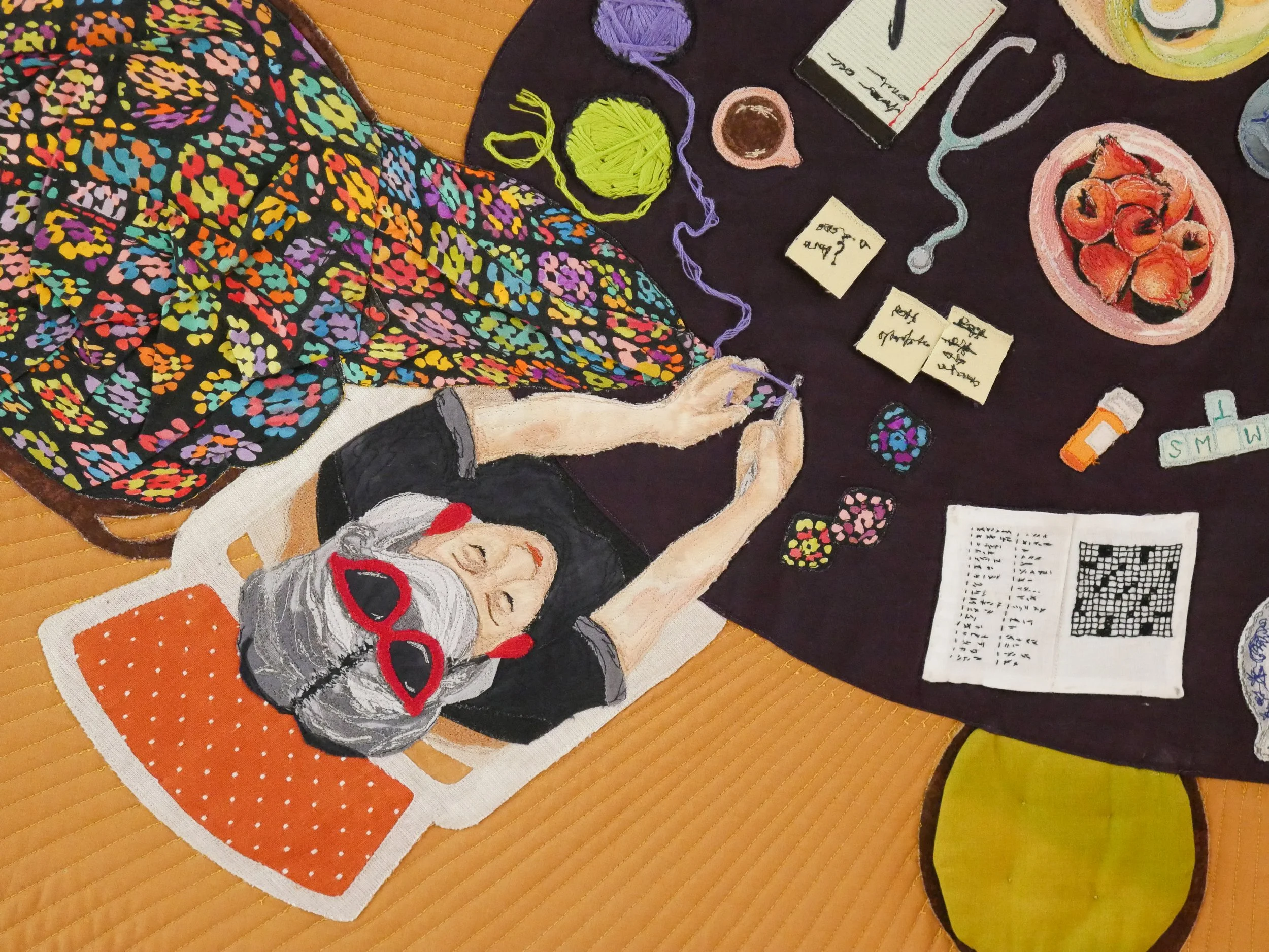 Quilt detail of a woman with grey hair sitting at a dinner table covered in trinkets and crocheting a large rainbow granny square afghan