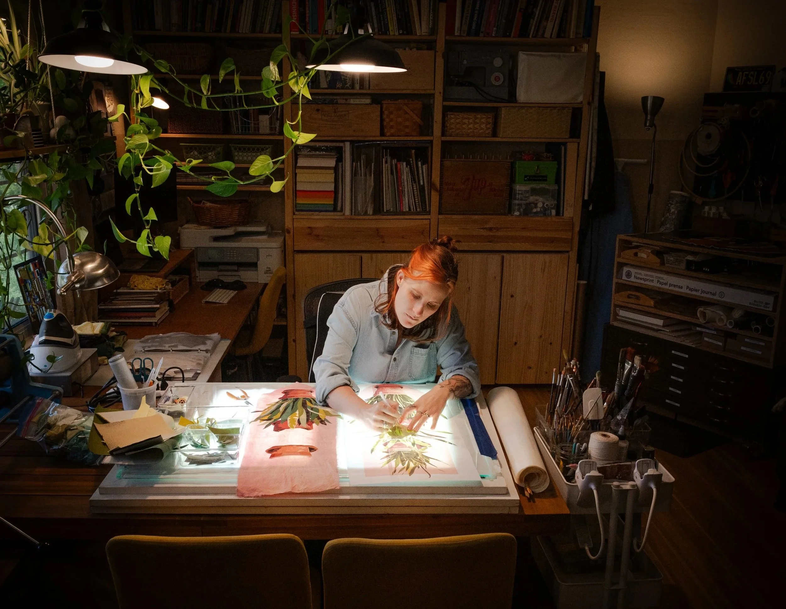 A white person with orange hair sits at a light table assembling an applique of a snake plant.