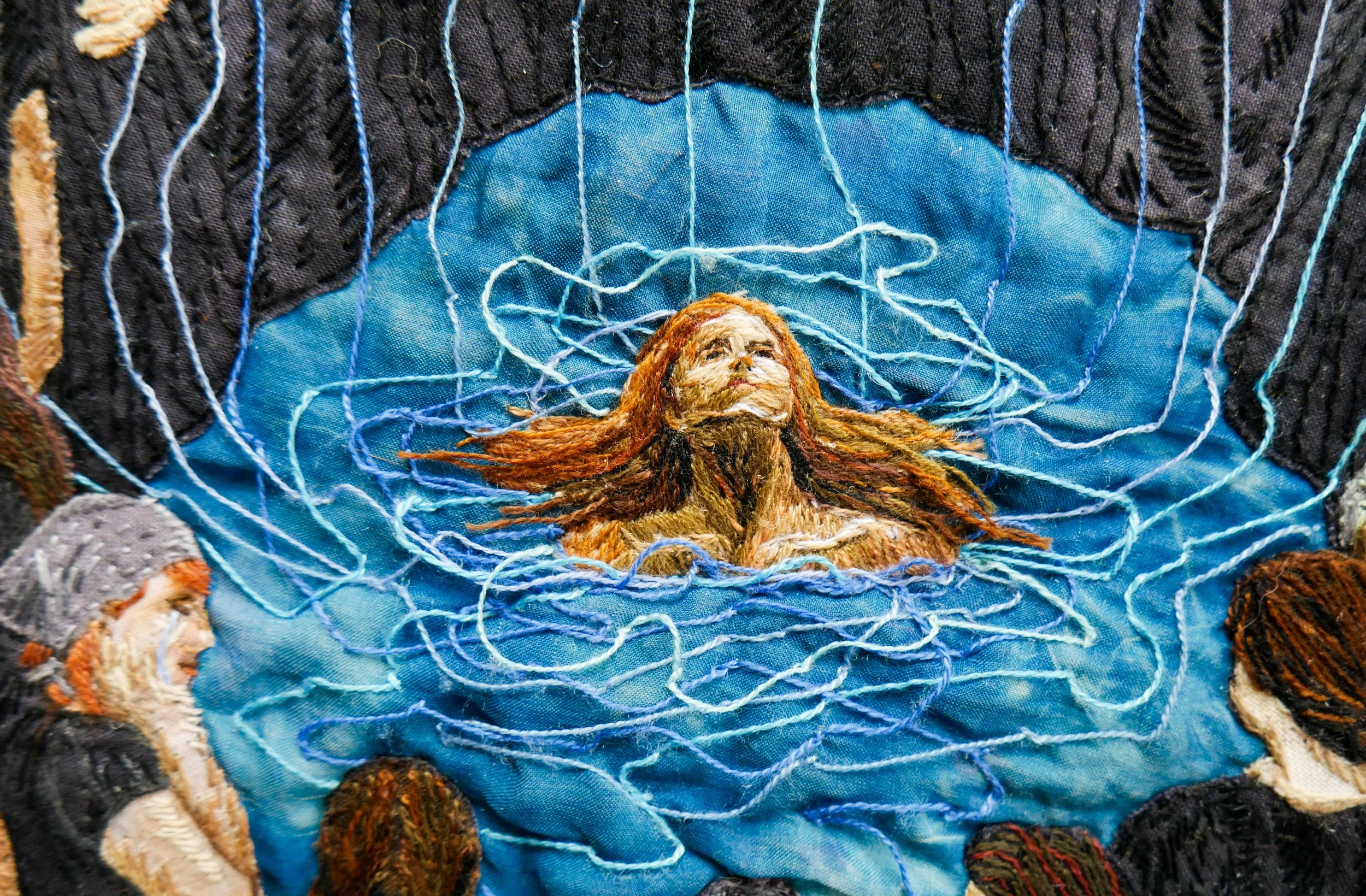 An embroidered face floats in blue water surrounded by blue threads