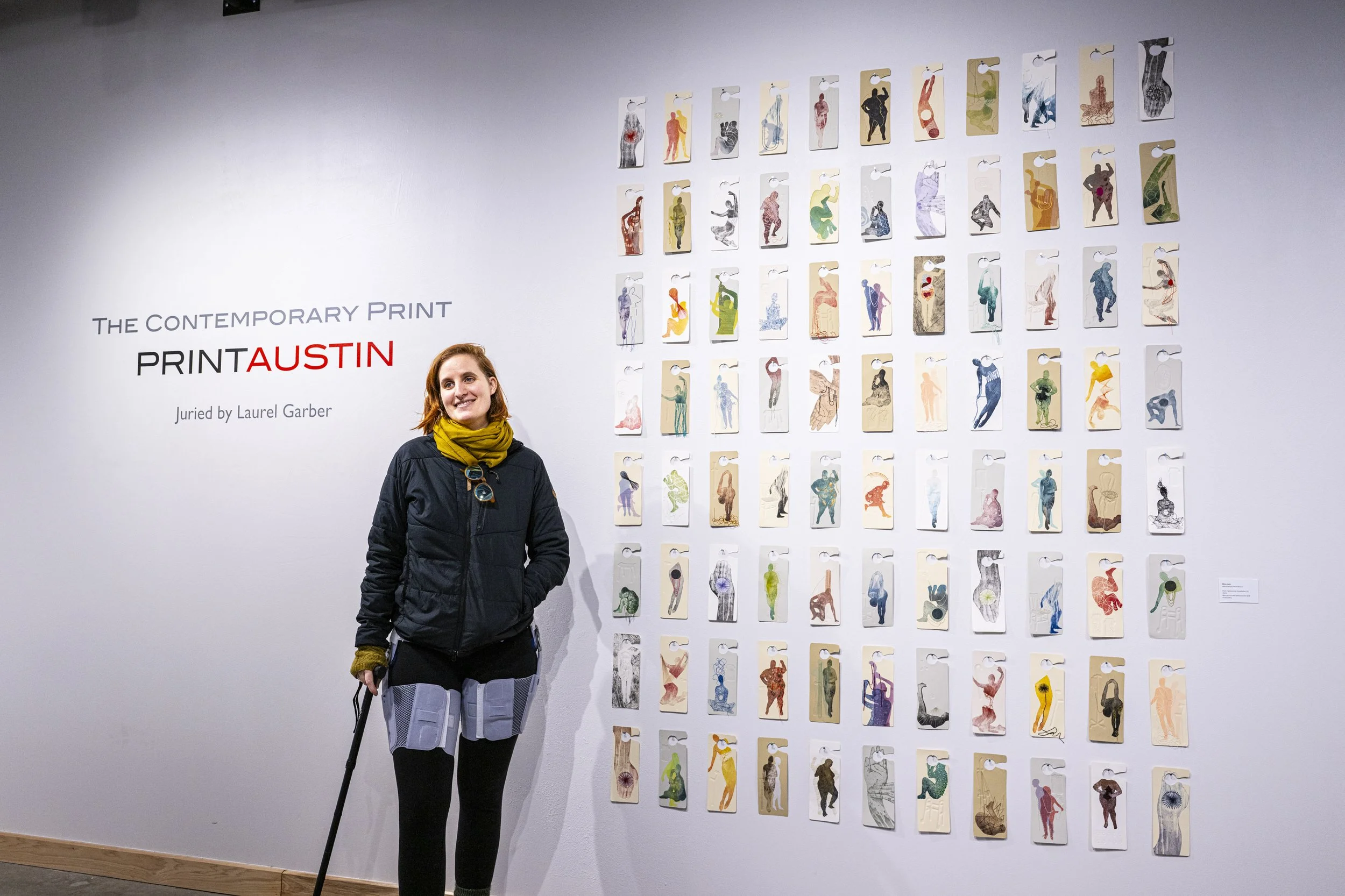 A non-binary person with cropped red hair wears a black puffy coat, a yellow scarf, and has a double hip brace on. They are standing with their black walking stick and smiling next to their print installation.