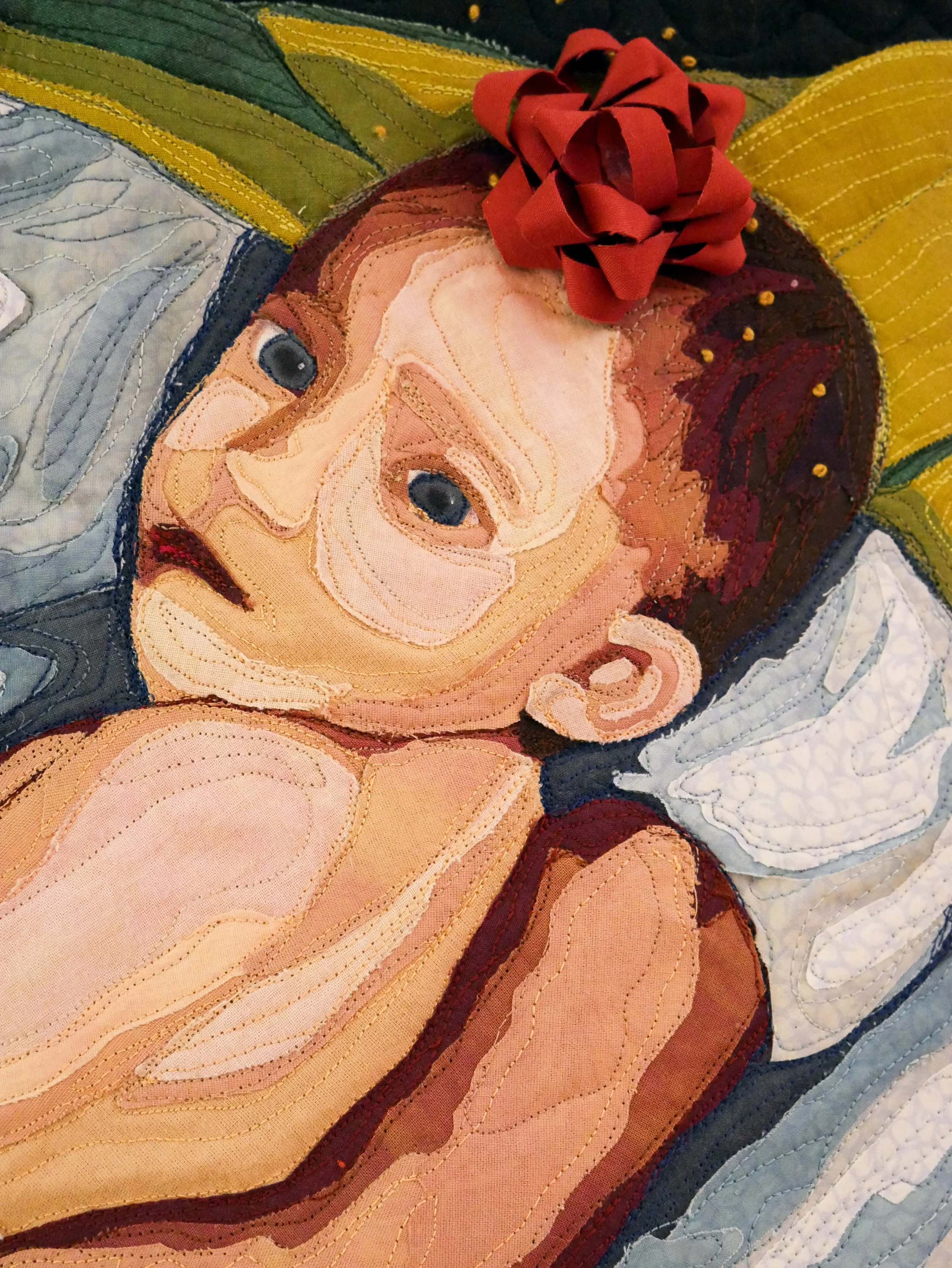 Quilt detail of the infant grandchild's face staring at the viewer with a gift wrapping bow on their head