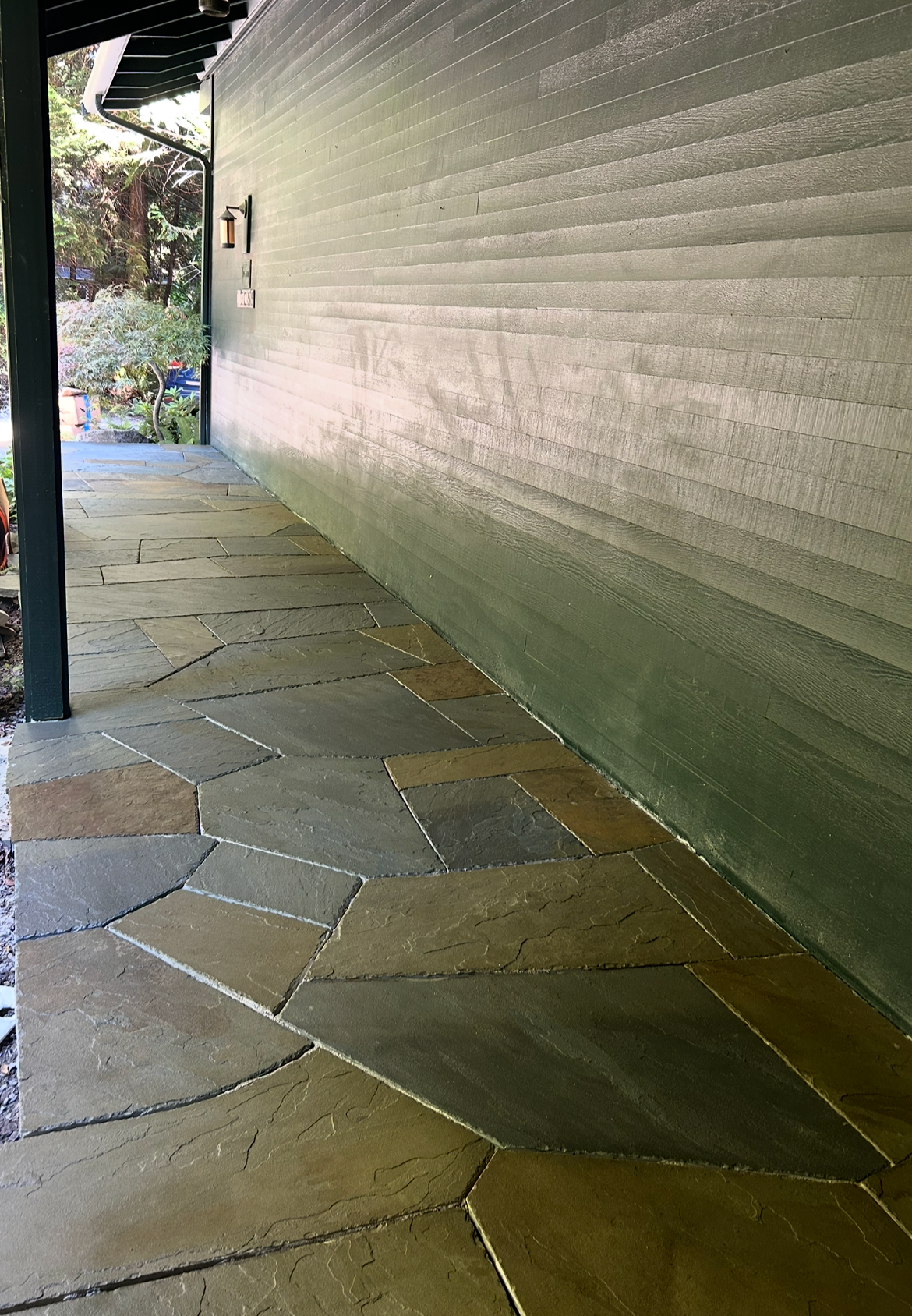 Image of a house exterior with a stone pathway and a green wooden wall. The pathway consists of irregularly shaped stone tiles, and there's a small garden area with trees and plants at the edge of the porch.