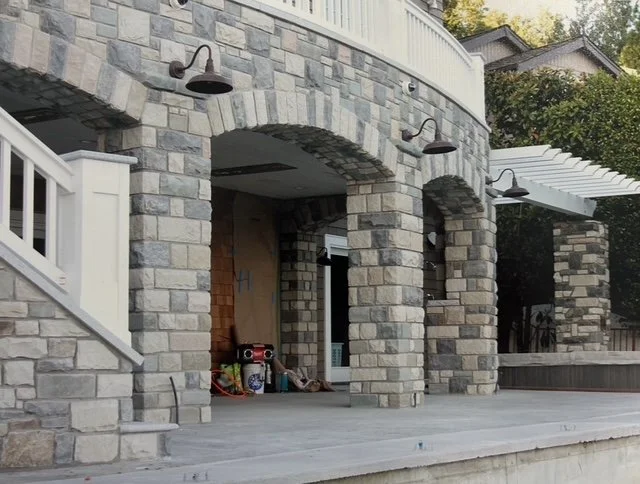A stone patio area with three arches supported by stone columns, with outdoor lights mounted on the stone facade, under construction or renovation, with tools and equipment on the ground. Wooden stairs with white railing lead up to the patio, and tre
