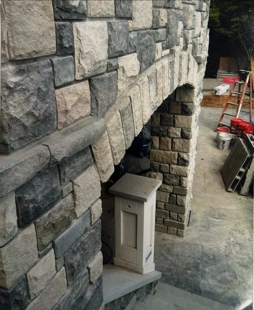 A stone fireplace with a wooden cabinet placed underneath it, under construction or renovation, with building tools and supplies on the ground nearby.
