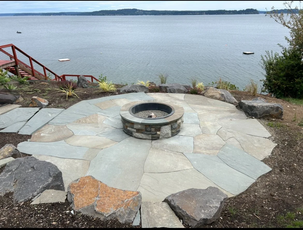 A circular stone fire pit built with stacked stones sits on a patio made of large, irregularly shaped flagstones overlooking a lake, with a red staircase leading down to the water on the left side and some small boats floating on the water.