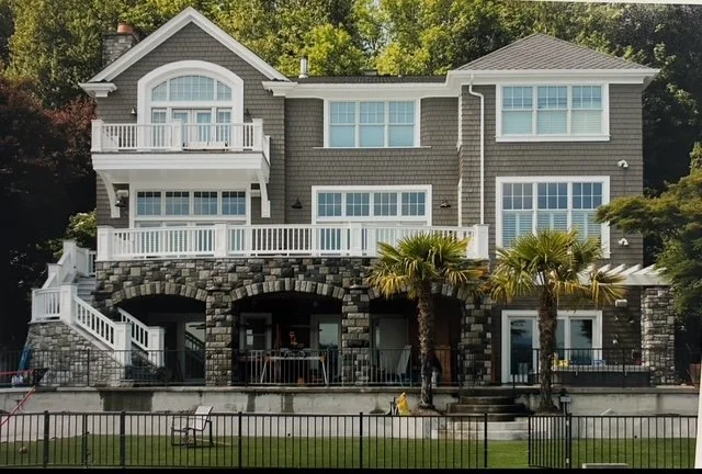 Large three-story house with gray exterior, multiple large windows, balconies, and a stone archway at the entrance. There are palm trees in the yard and an iron fence in the foreground.