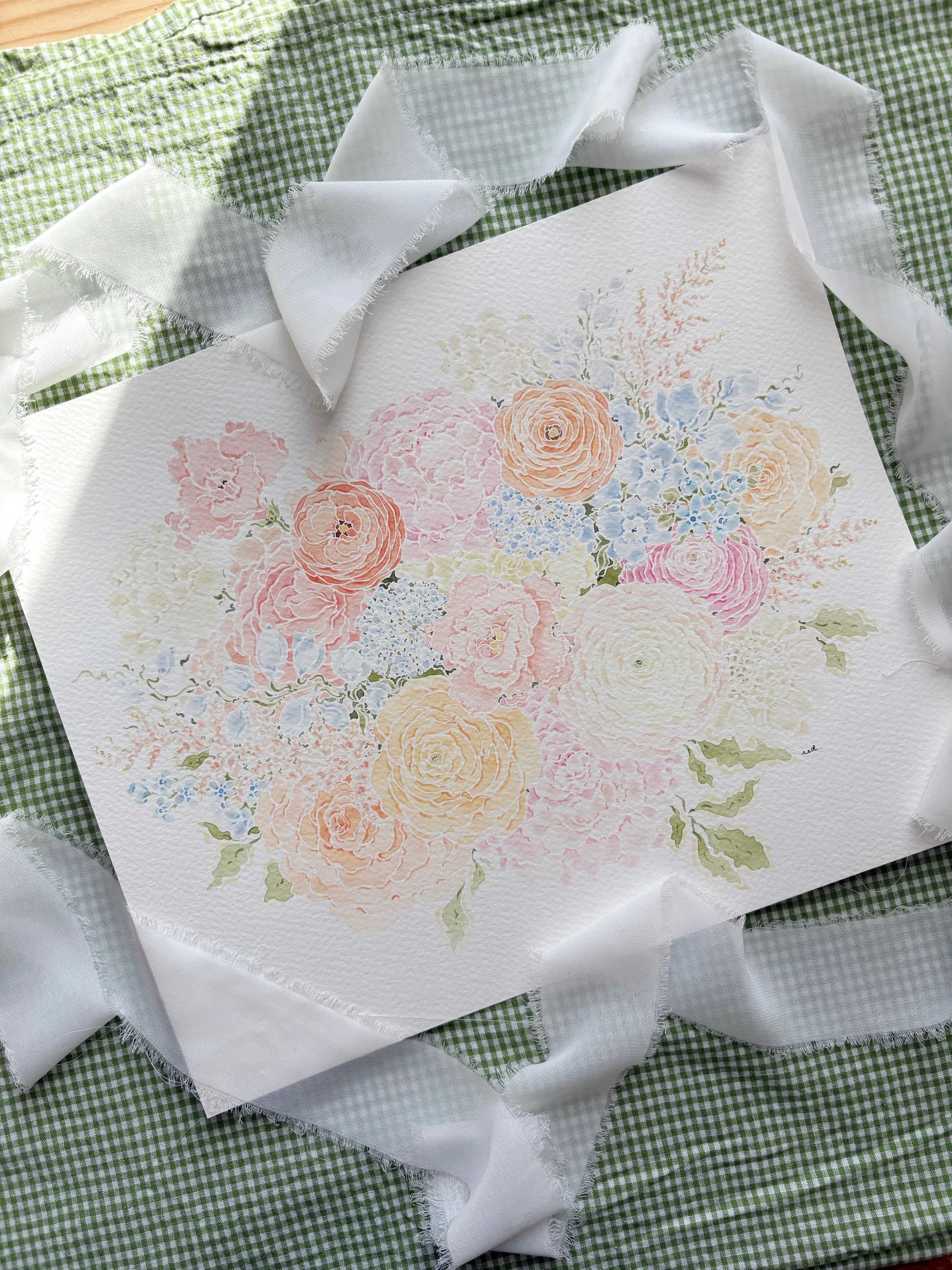 A watercolor painting of pastel-colored flowers, including roses and smaller blossoms, on textured paper, surrounded by white fabric ribbons on a green checkered tablecloth.