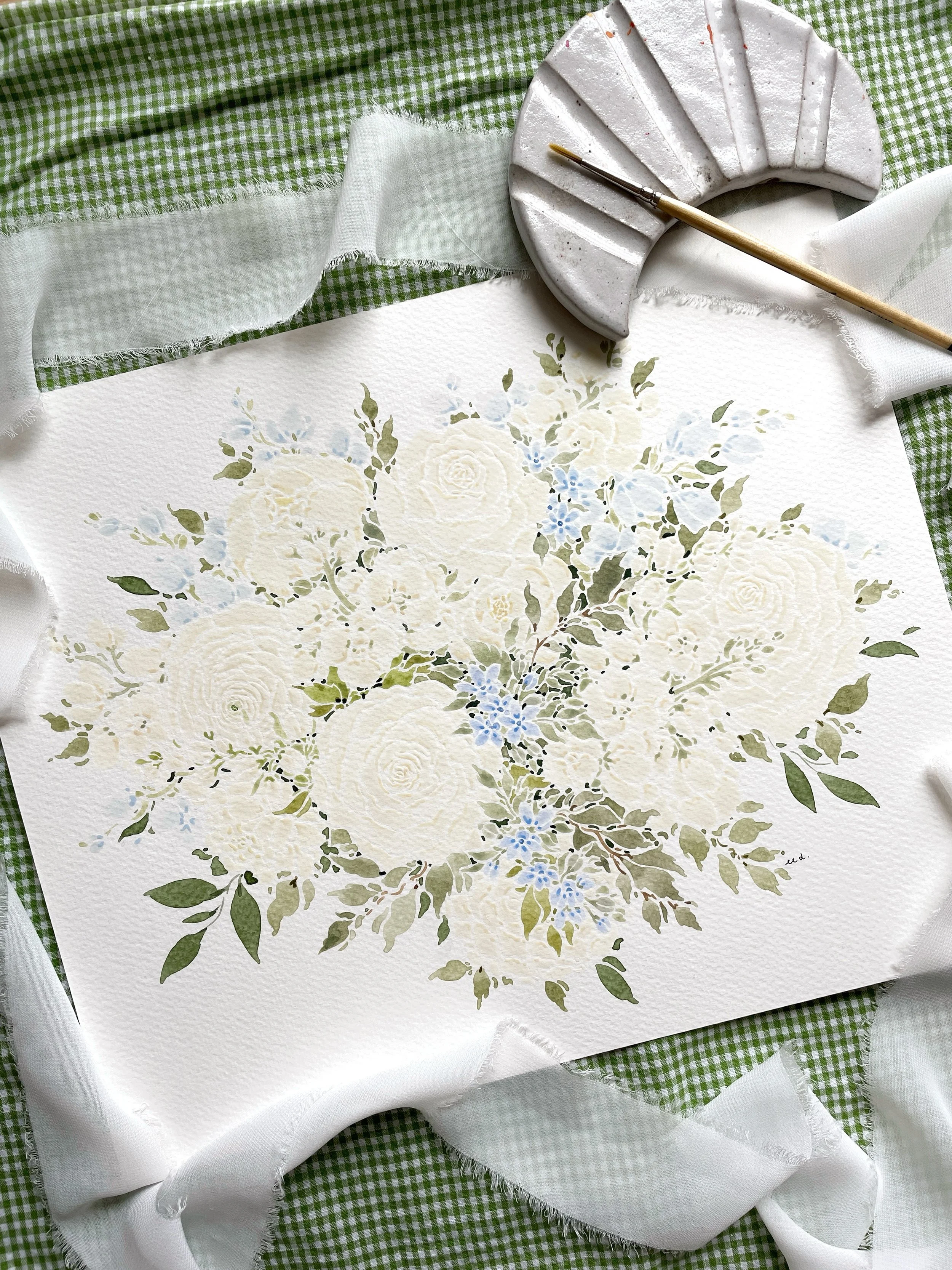 A watercolor floral painting featuring white roses, green leaves, and small blue flowers, on textured paper, surrounded by a green checkered fabric, lace, a white cloth, and an artist's brush.